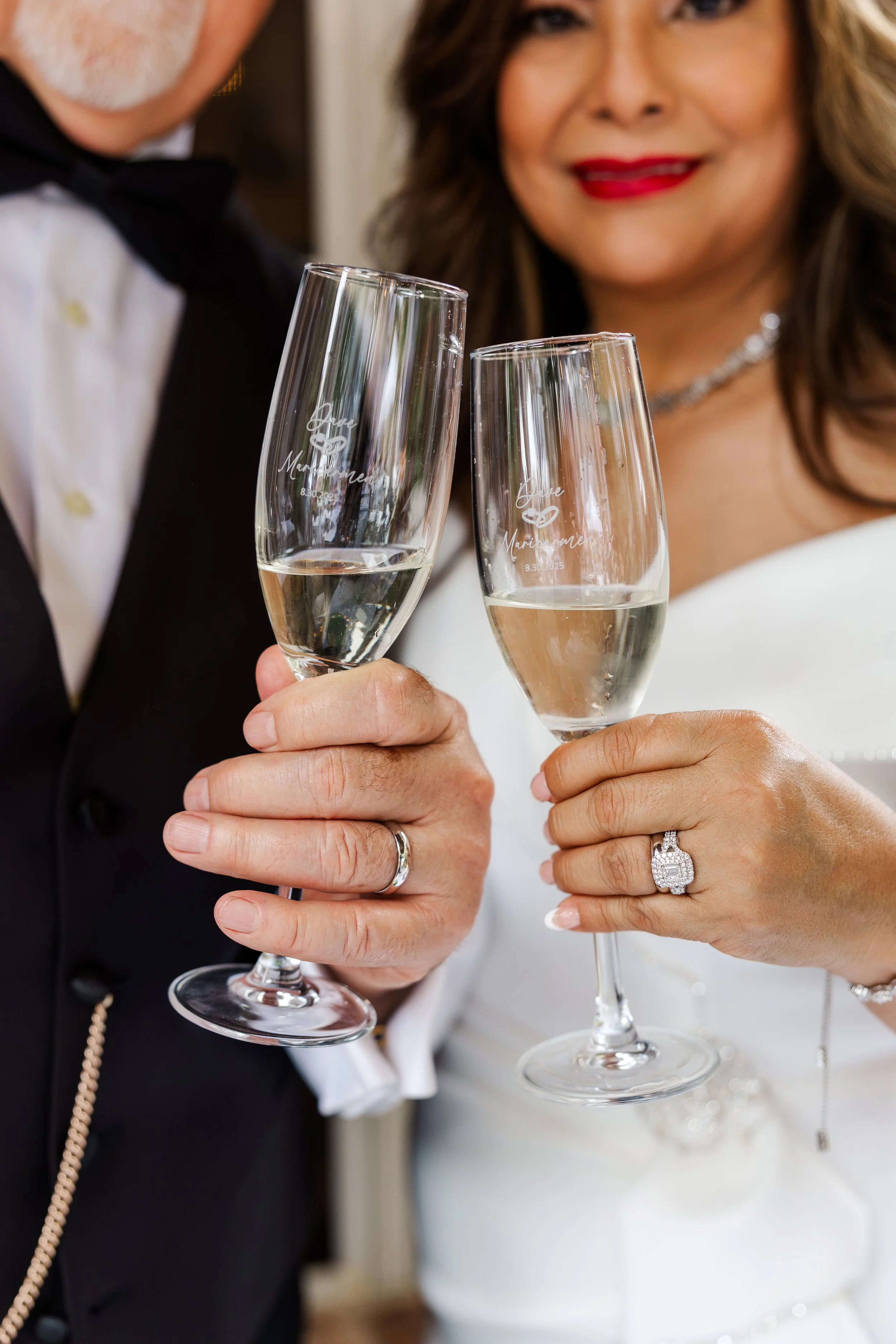 Close-up of a newlywed couple holding glasses of champagne, the bride wearing a wedding ring and the groom wearing a wedding ring, with the bride smiling.