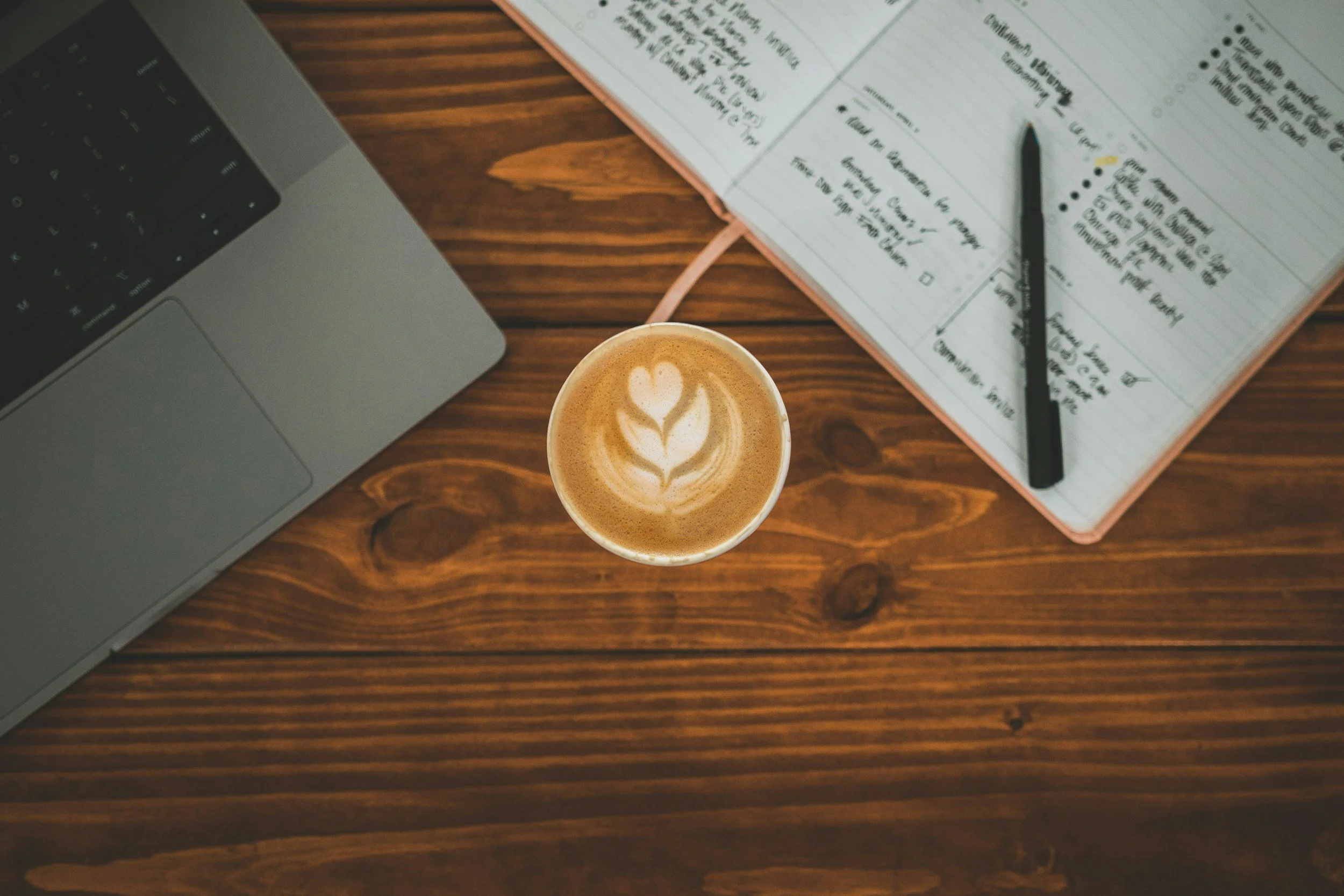 Overhead view of a wooden table with a laptop, a notebook with handwritten notes, a pen, and a cup of coffee with latte art.