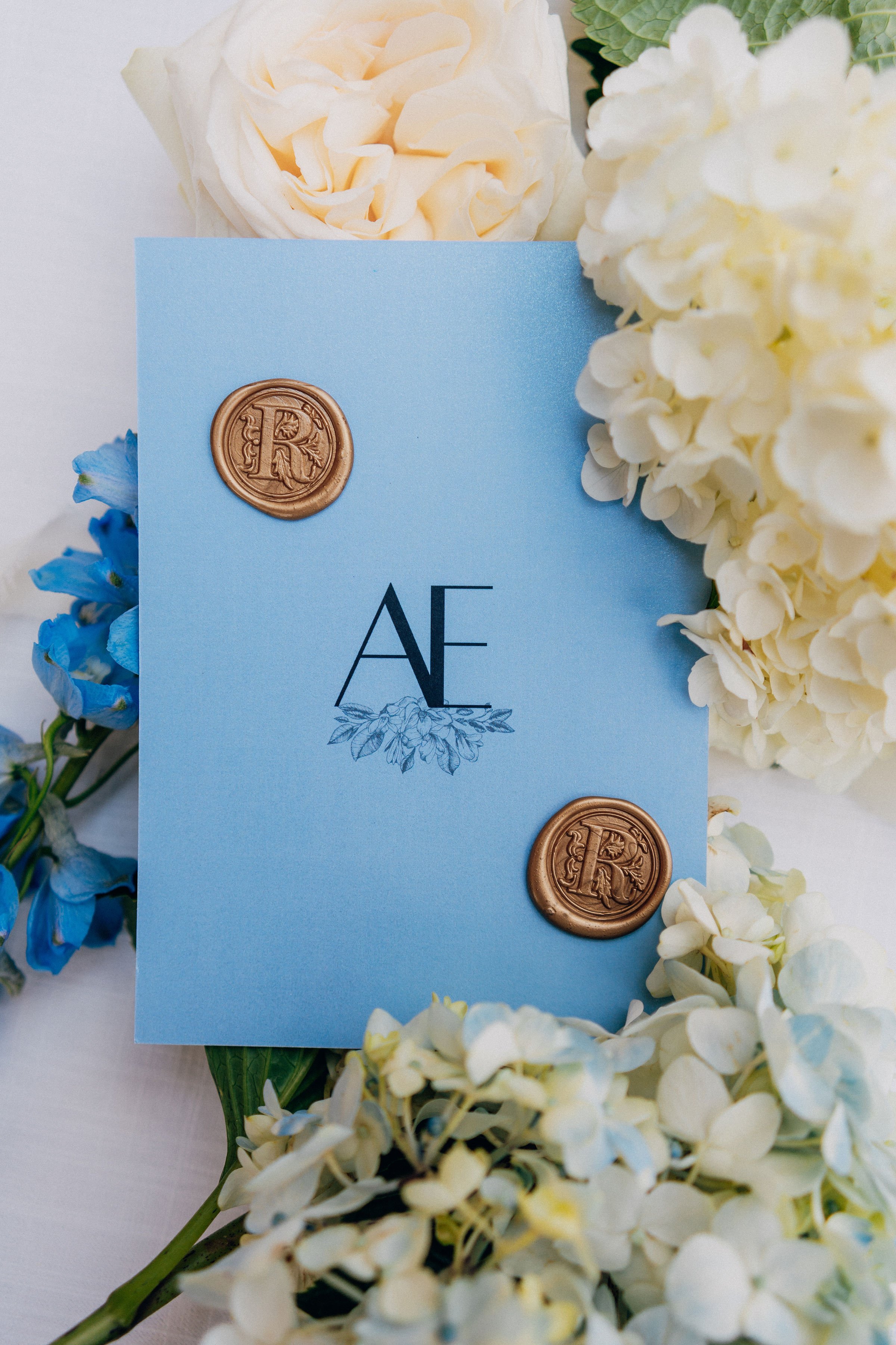 A light blue wedding invitation card with black initials 'AE', decorated with a floral illustration, sealed with two gold wax seals, surrounded by white, cream, and blue flowers.