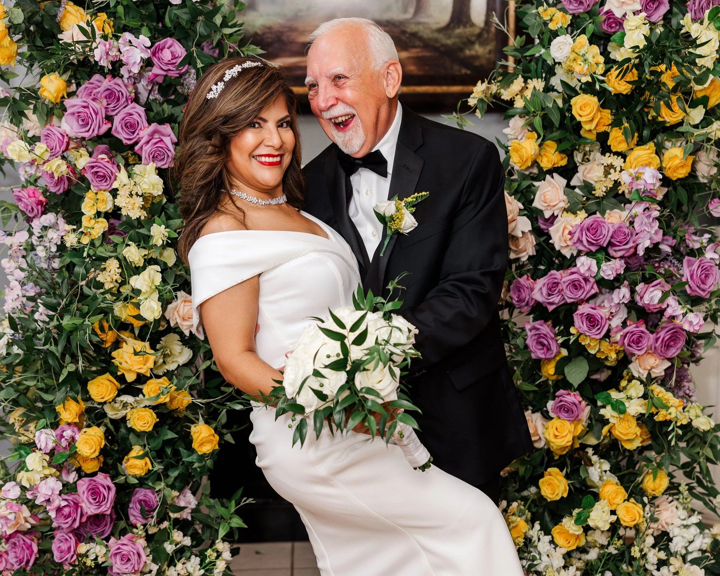 A woman in a white wedding dress holding a bouquet of white roses, standing beside a man in a black tuxedo with a boutonniere, against a backdrop of colorful pink, yellow, and purple roses and greenery at a wedding celebration.