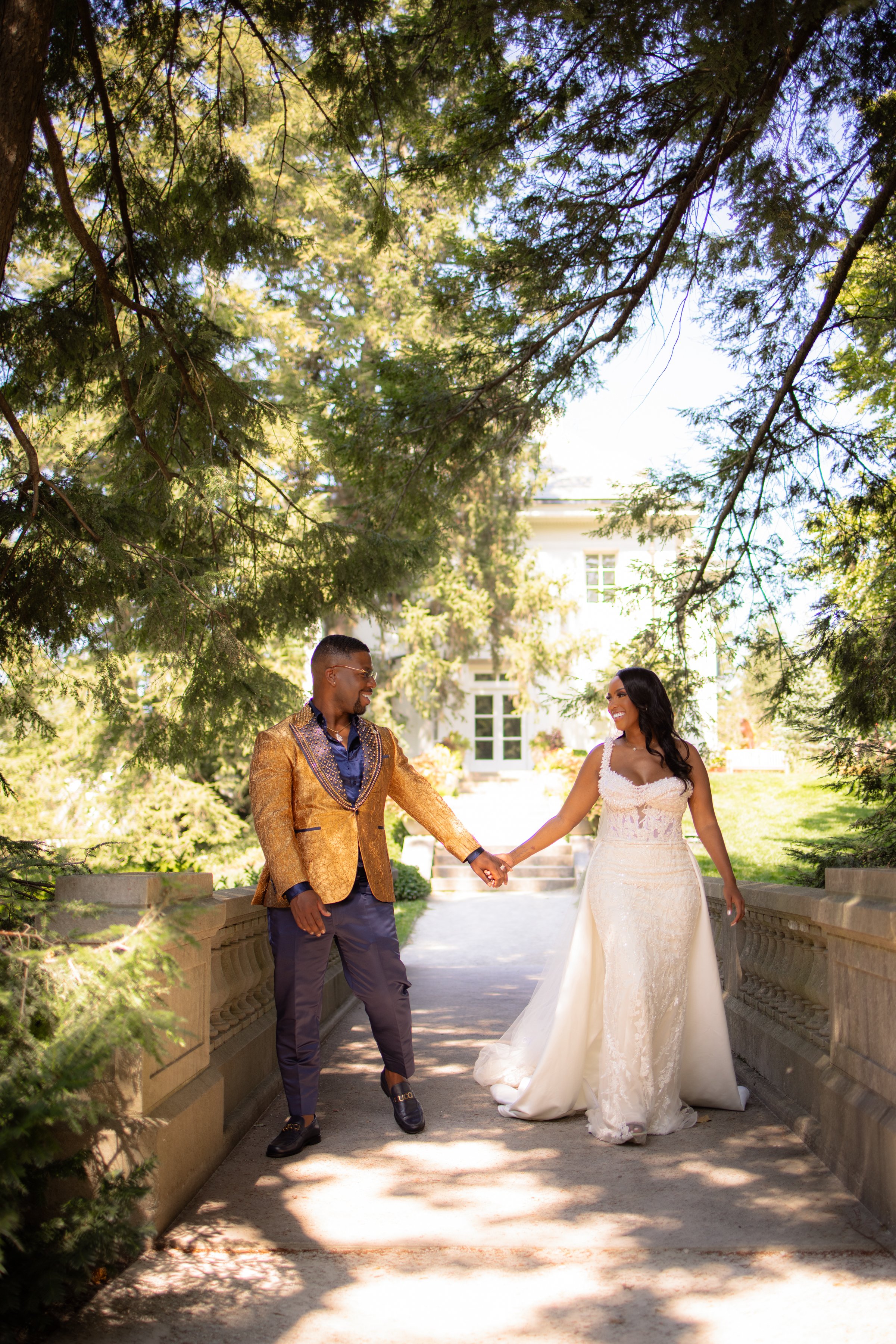 A newlywed couple holding hands and smiling at each other on a bridge surrounded by green trees in a garden.