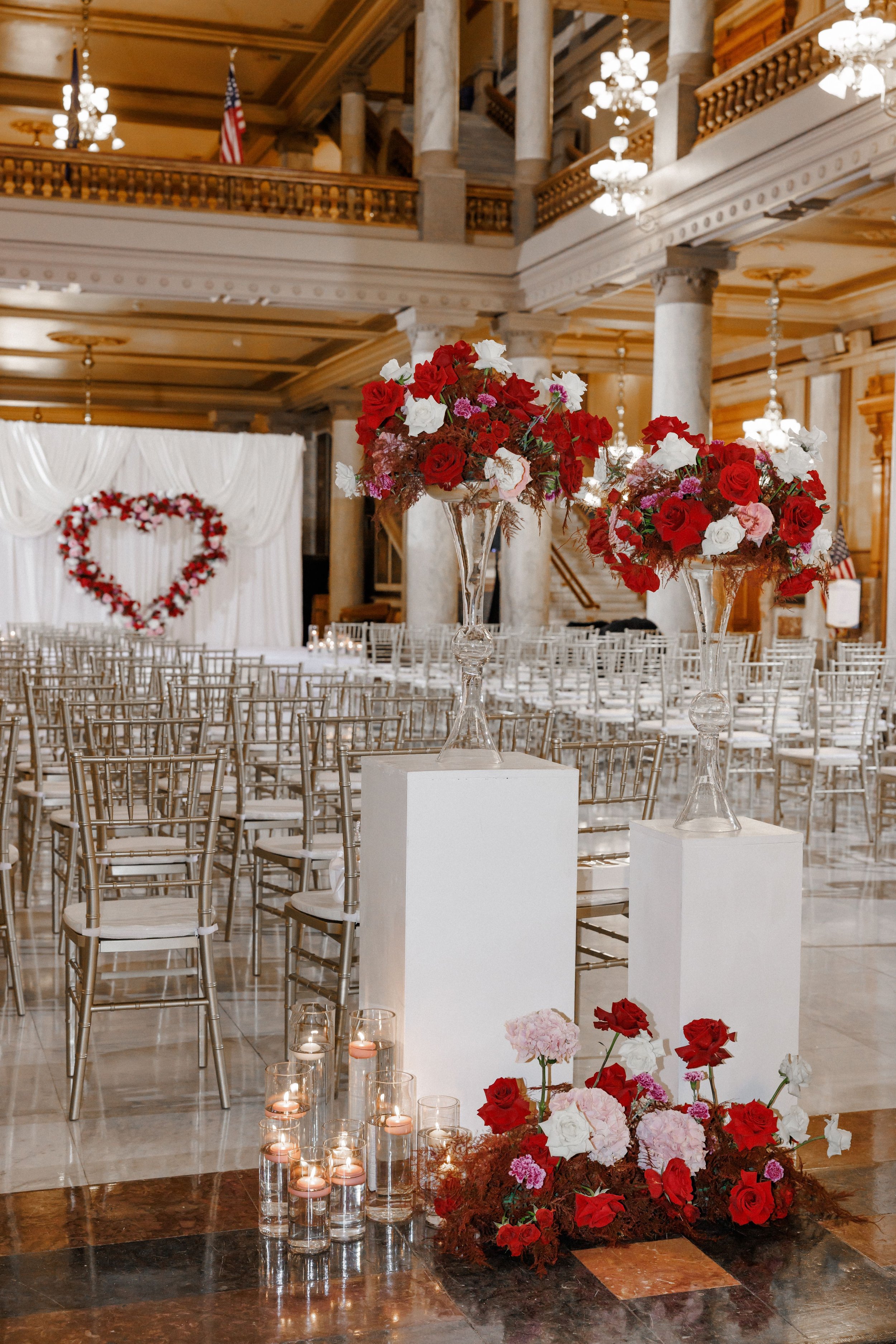 Elegant wedding ceremony setup with flower arrangements on white pedestals, candles in glass holders, and a heart-shaped floral backdrop in the background inside a grand hall with tall columns and chandeliers.