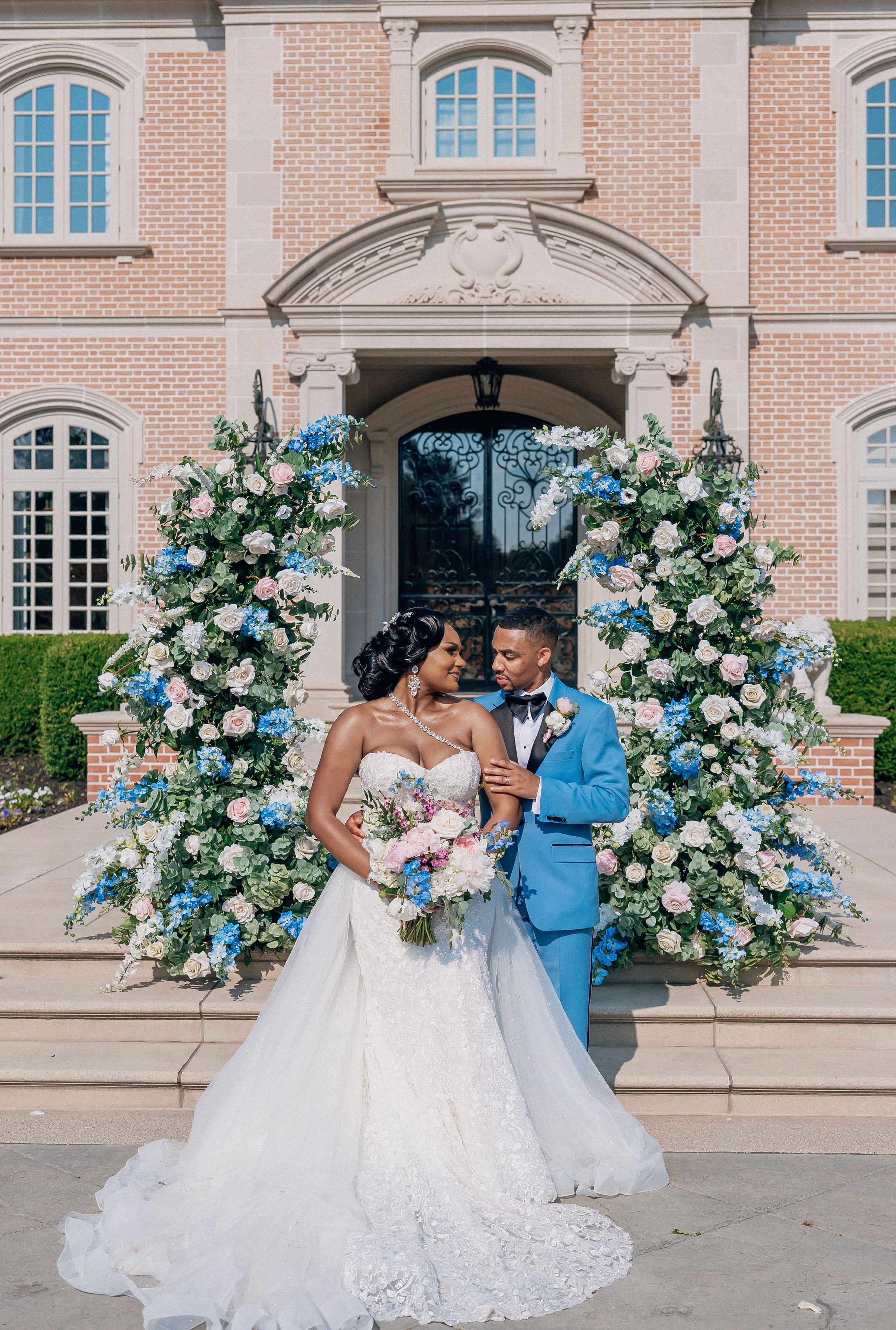 A bride and groom standing in front of a pink brick mansion decorated with large floral arrangements, with the bride holding a bouquet and wearing a white wedding gown, and the groom in a blue tuxedo.