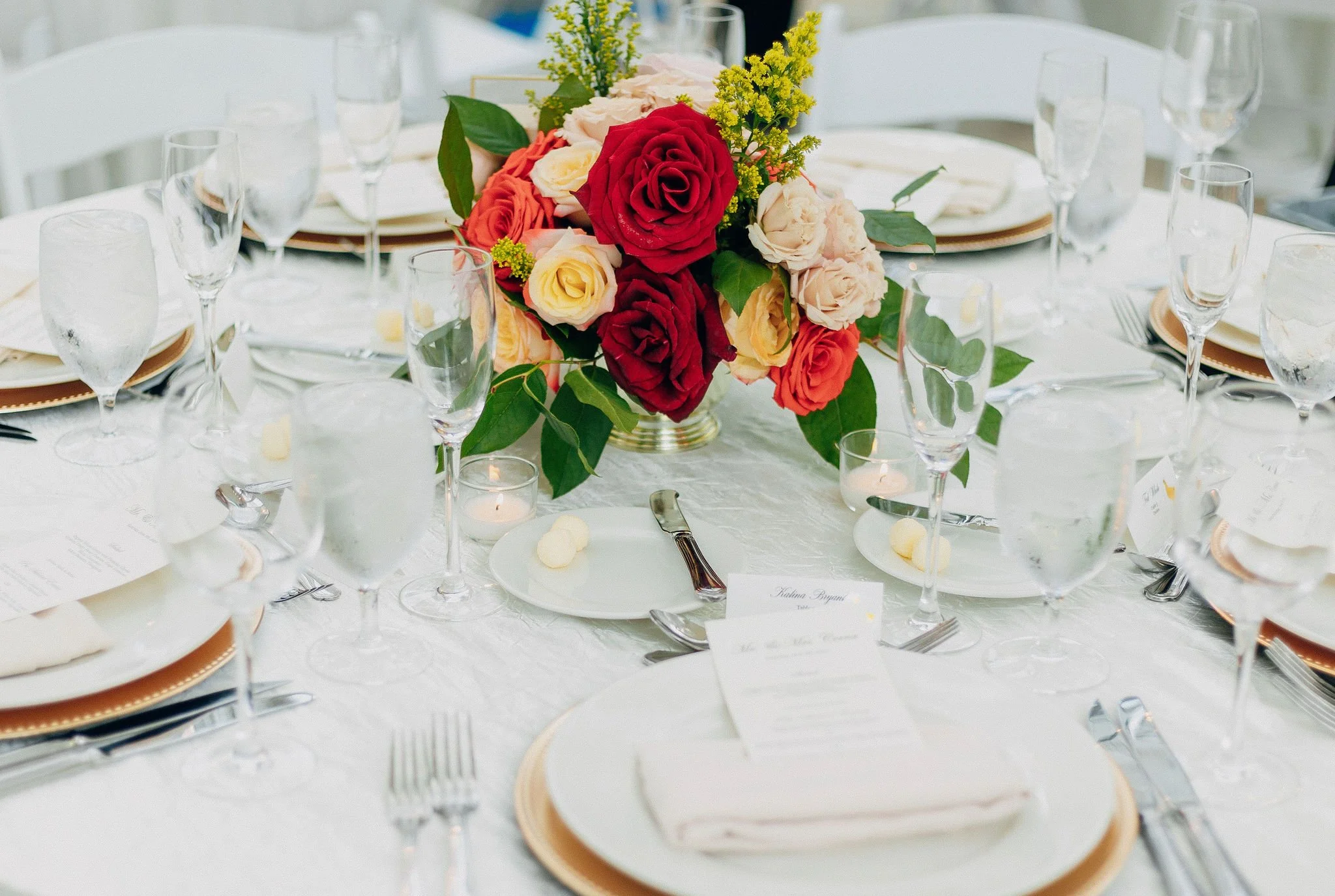 Elegant wedding table setting with a floral centerpiece of red, cream, and yellow roses, surrounded by glassware, plates, silverware, and small candles on a white tablecloth.