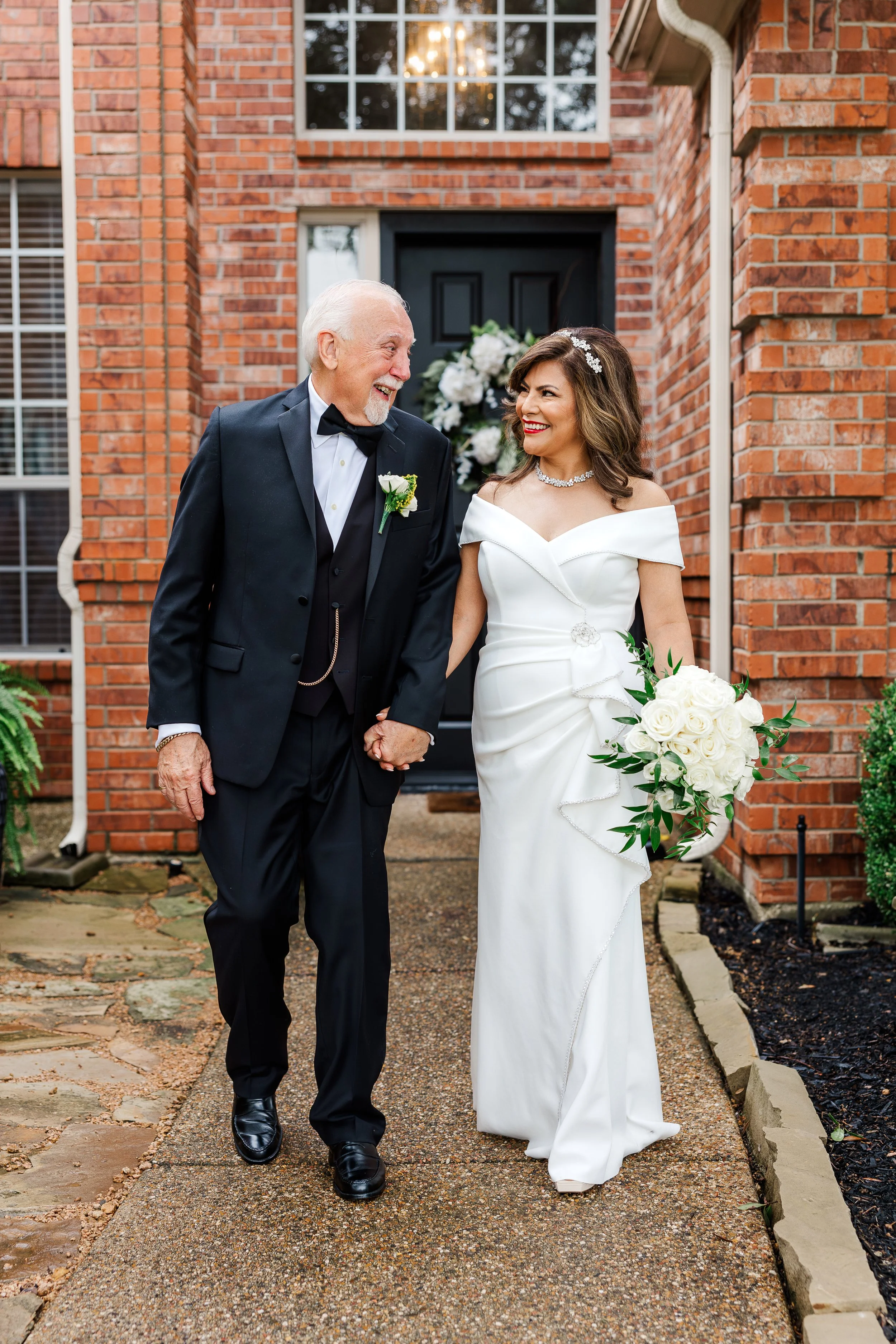 A bride in a white wedding dress holding a bouquet of white roses, walking hand-in-hand with an older man in a black tuxedo in front of a brick house.