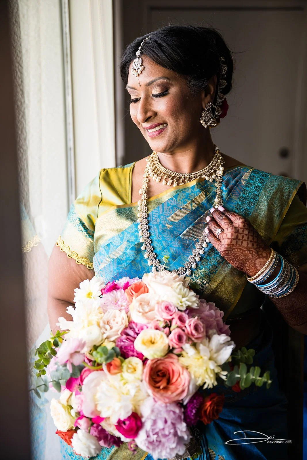 A smiling woman wearing traditional Indian attire and jewelry, holding a bouquet of colorful flowers, standing near a window.
