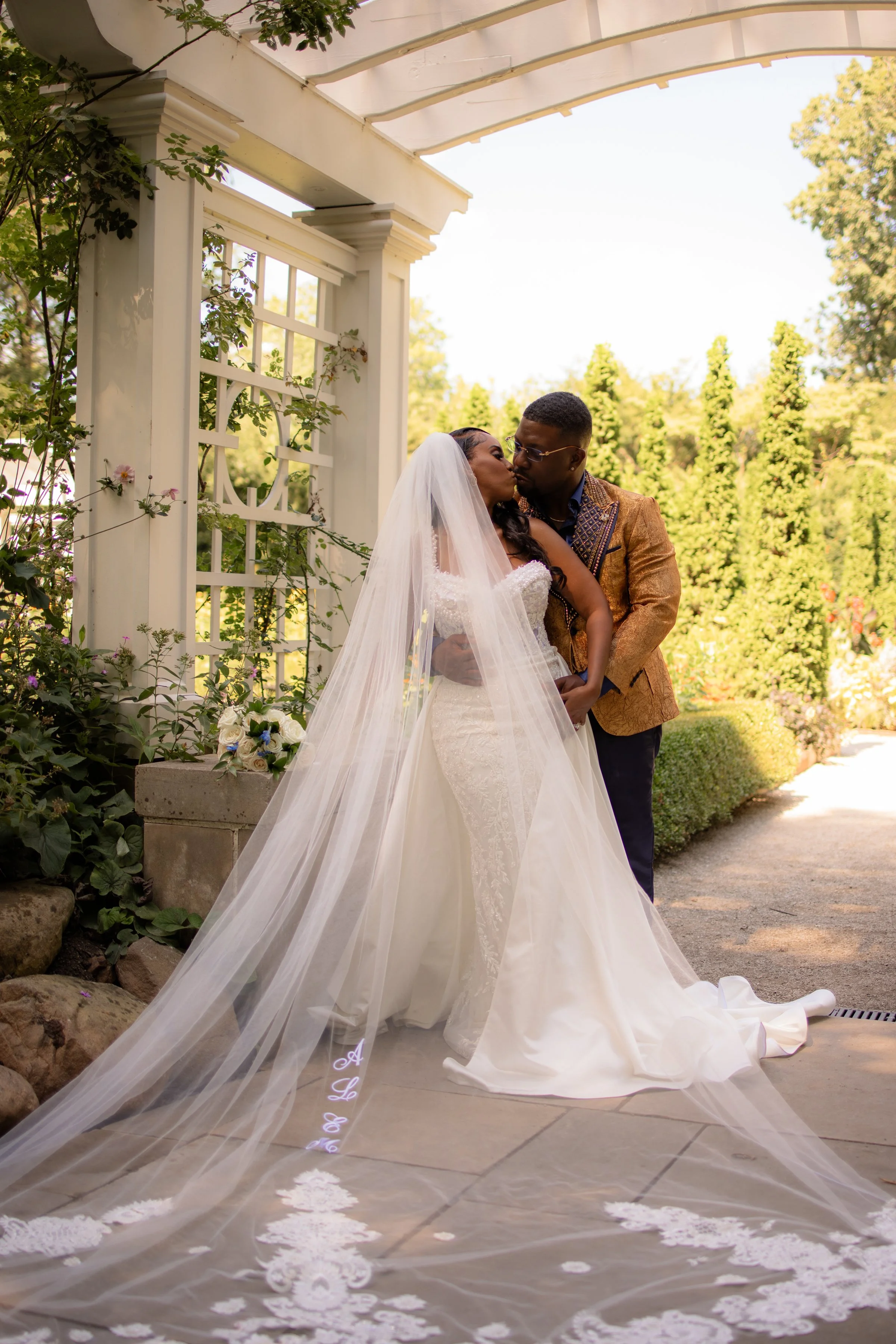 A bride and groom sharing a kiss in an outdoor garden setting, with the bride in a white wedding gown and veil, and the groom in a brown suit jacket.