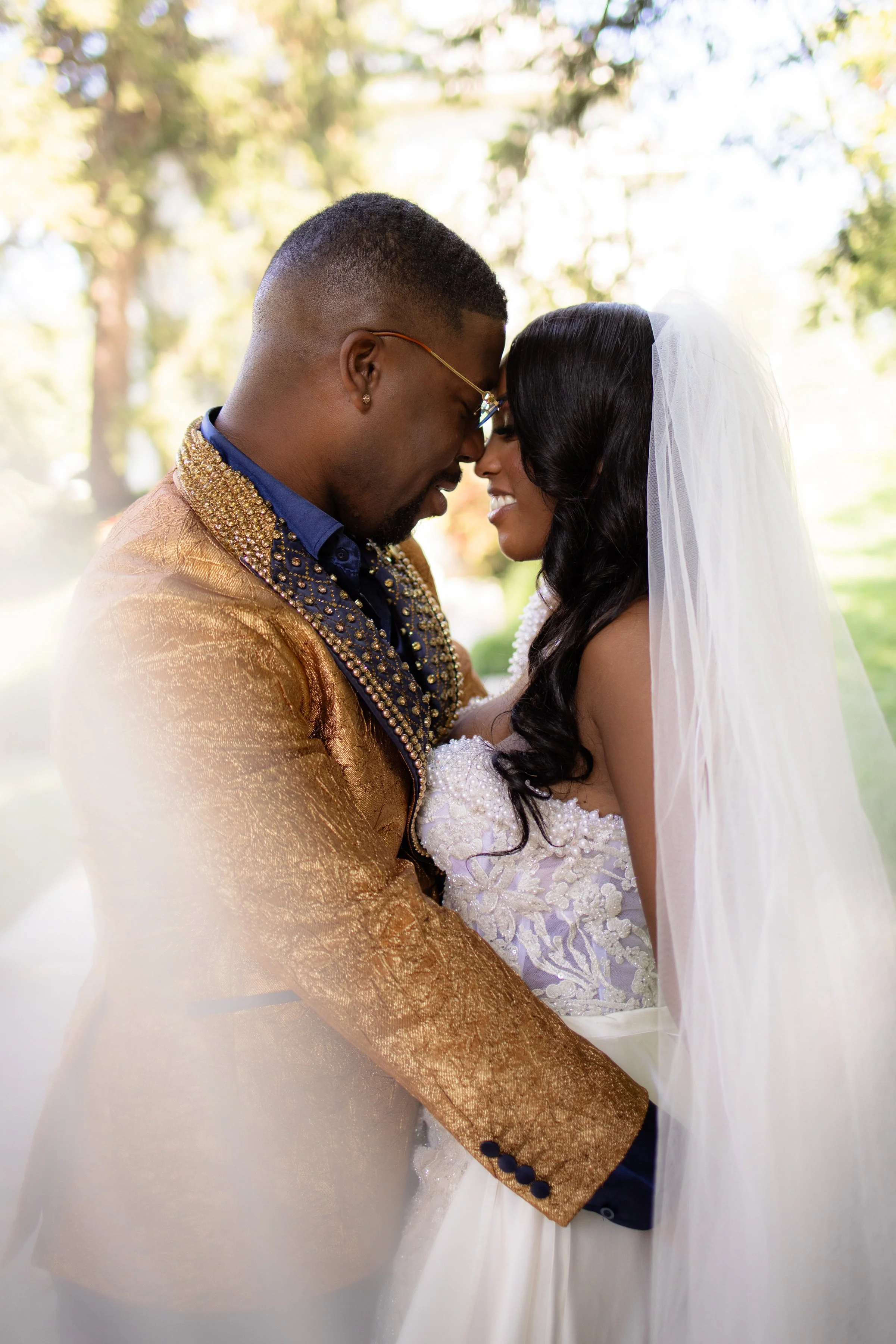 A bride and groom are holding each other with their foreheads touching, smiling outdoors during their wedding.