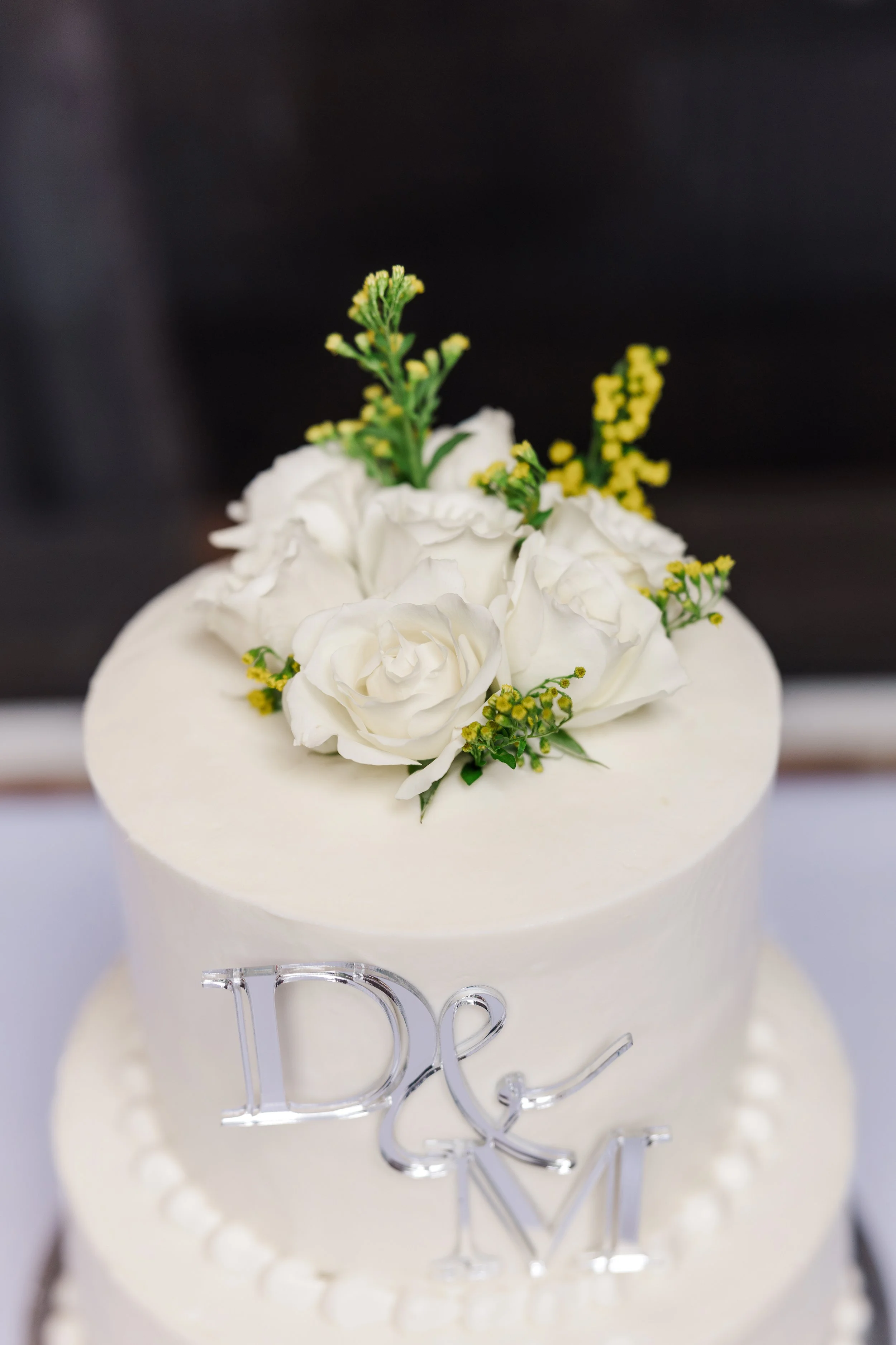 A white wedding cake decorated with real white roses and yellow-green flowers, with a silver 'D & M' cake topper.