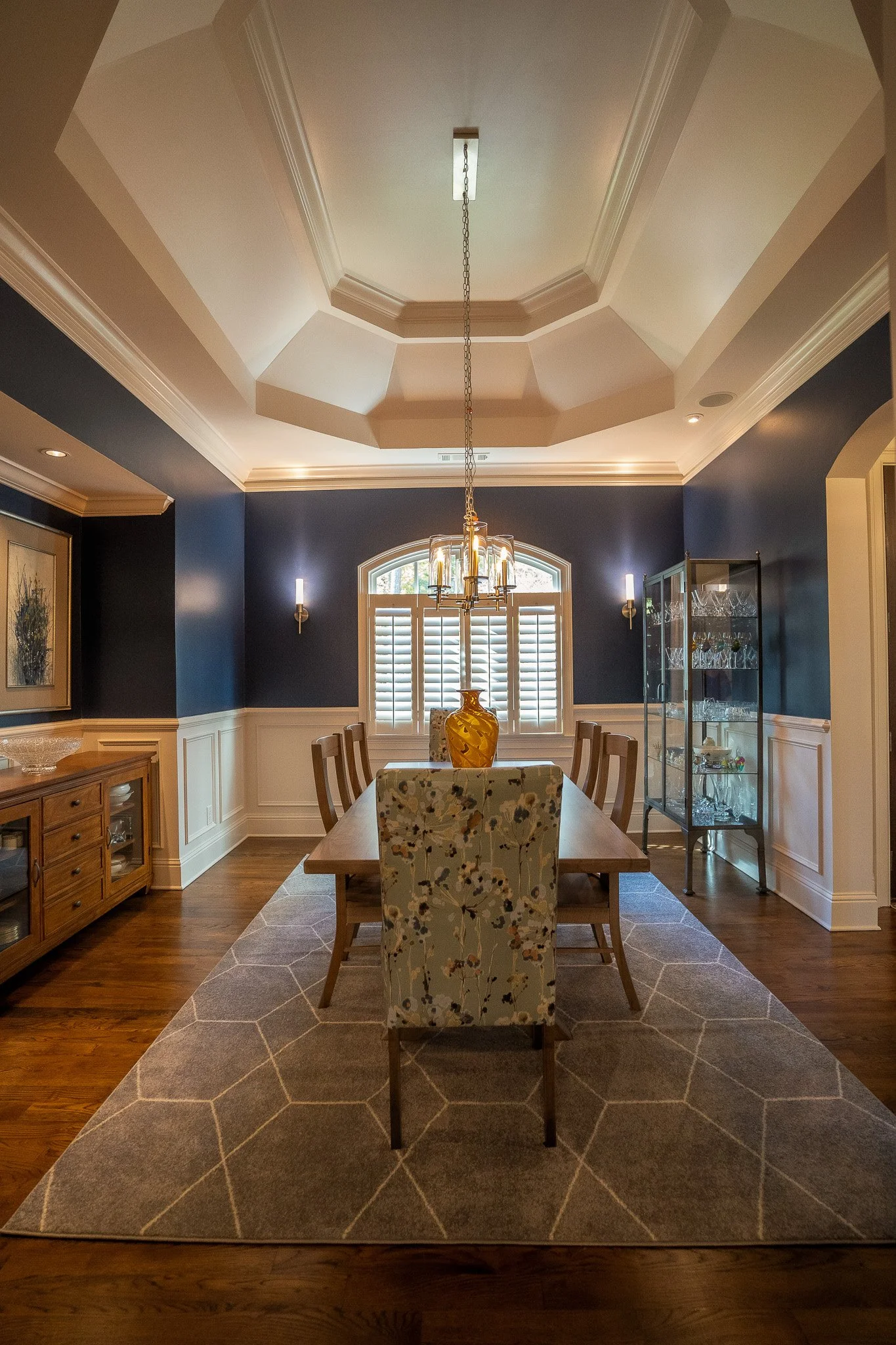 A dining room with two-tone blue walls, white wainscoting, and a wooden dining table with six chairs. A large, light gray area rug with a hexagonal pattern covers the hardwood floor. A chandelier hangs above the table, and there's a window with plantation shutters on the back wall. The room has decorative wall sconces and a glass display cabinet on the right side.