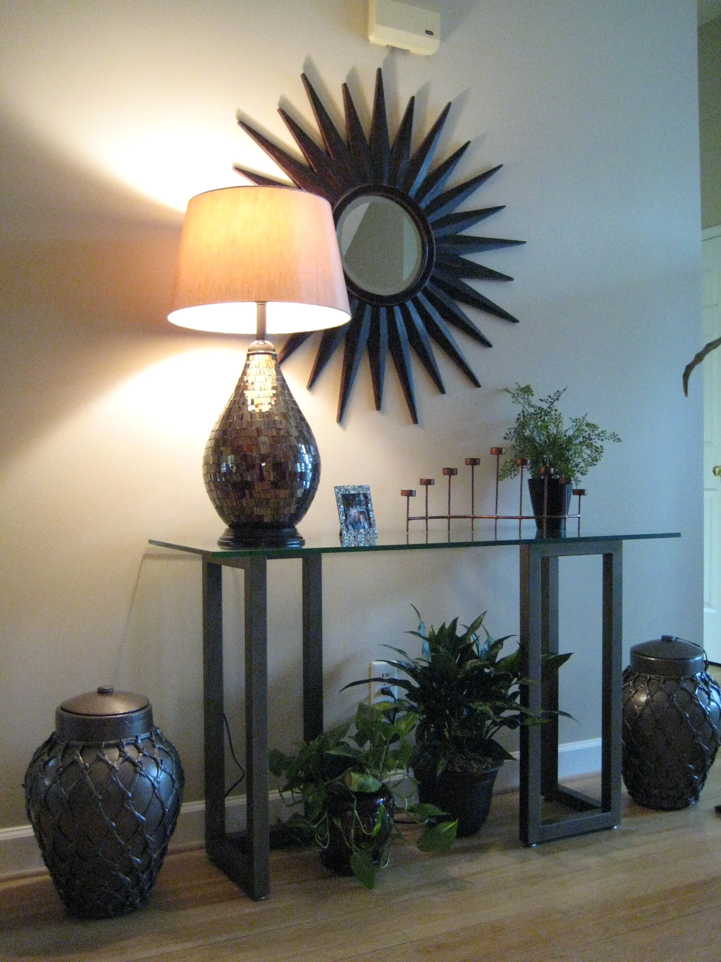 Warm entryway styling featuring a sculptural sunburst mirror, glass console table, ambient table lamp lighting, and layered greenery creating a cozy transitional interior design atmosphere.