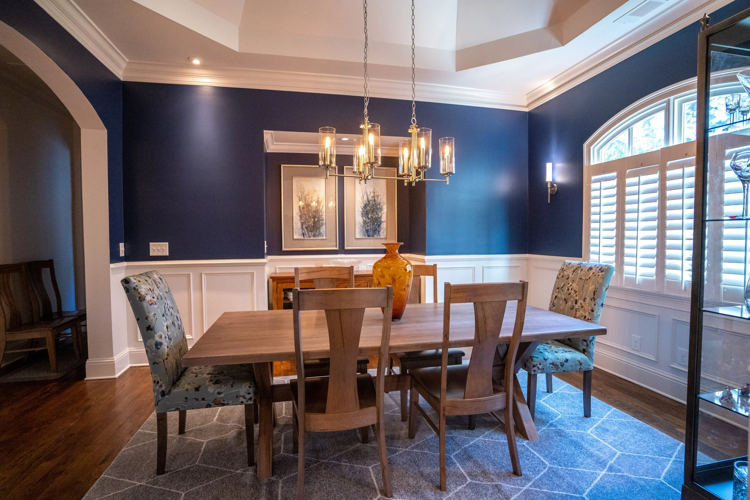 Elegant dining room featuring deep navy blue walls, white millwork, warm wood flooring, modern chandelier lighting, and a natural wood dining table with upholstered chairs in a timeless transitional interior design.