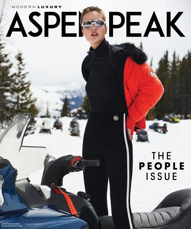 Woman standing next to a snowmobile in a snowy mountain landscape, wearing sunglasses, a black turtleneck, and a red jacket, with trees and snow-covered mountains in the background.