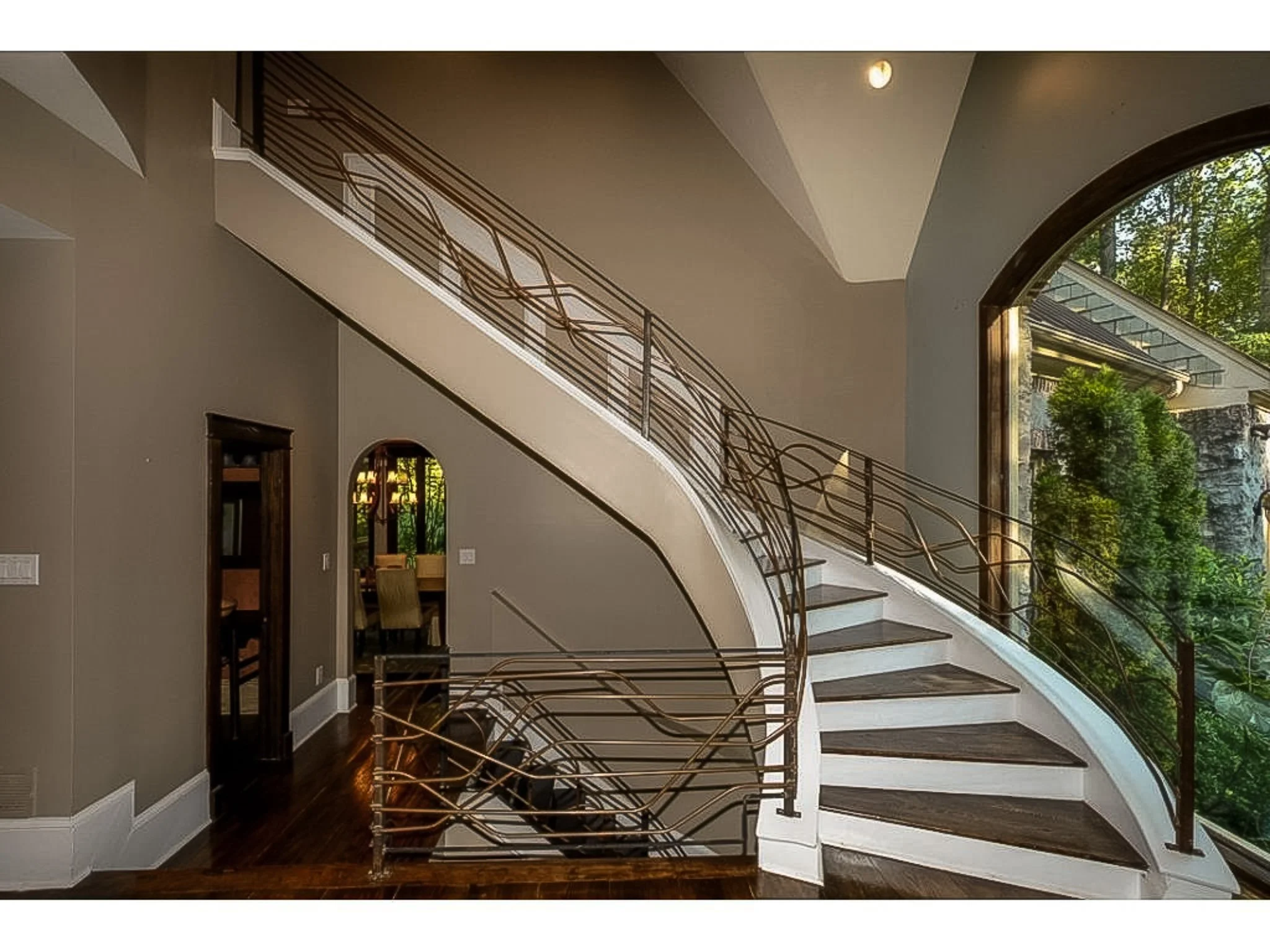 Interior of a modern home with a staircase featuring decorative metal railings and large arched windows showing trees outside.