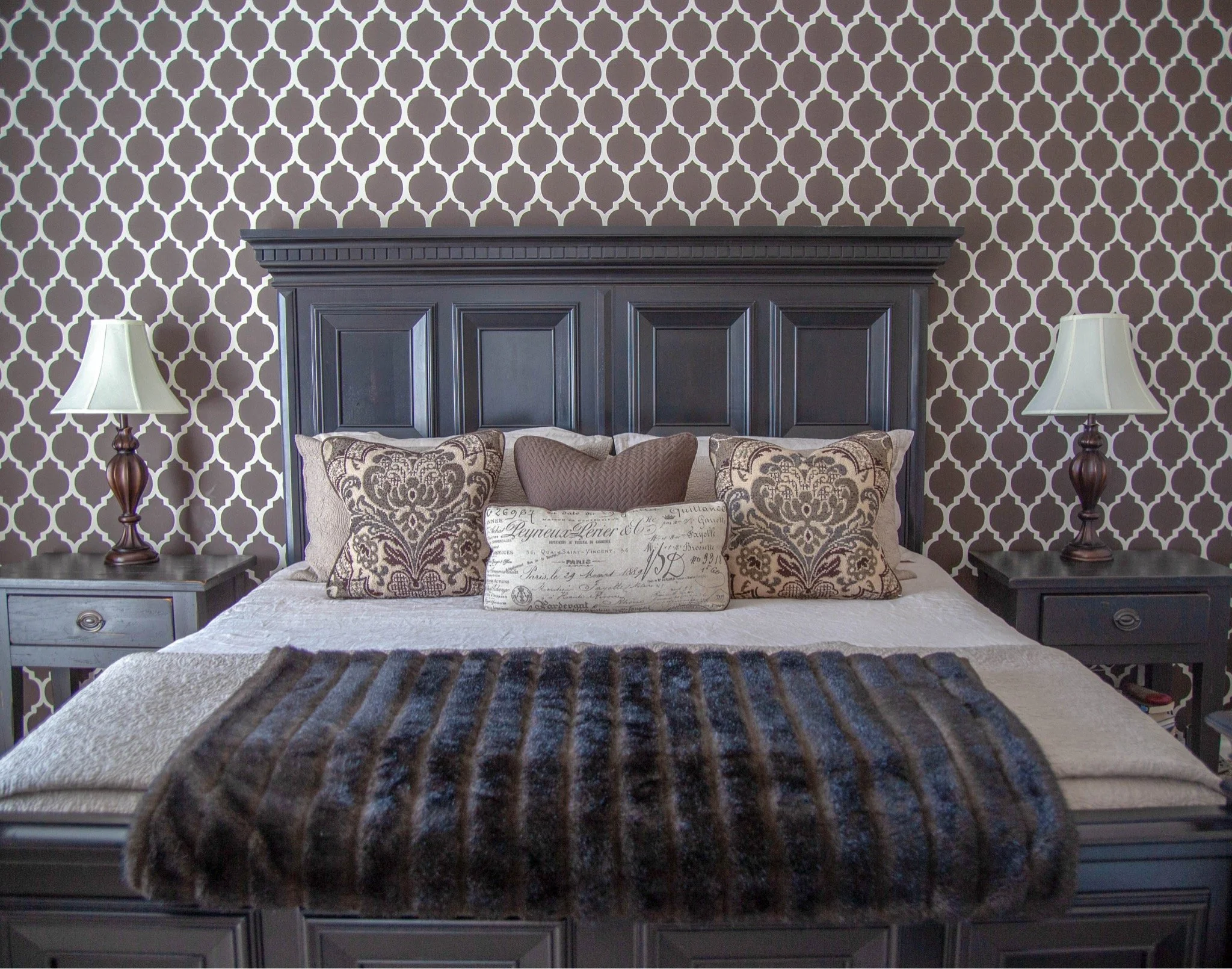 A bedroom with a black wooden headboard, patterned wallpaper, and two matching lamps on bedside tables. Cushions with ornate and handwritten designs are placed on the bed.