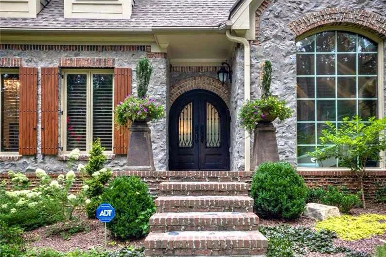French Country stone home exterior with arched iron front doors, brick steps, oversized windows, wood shutters, and lush landscaping designed with timeless European-inspired architecture.