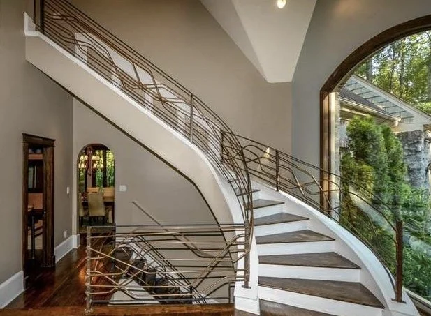 Elegant curved staircase featuring custom copper railing, warm wood flooring, and oversized arched windows in a timeless luxury interior designed by Velvet Oak Home.