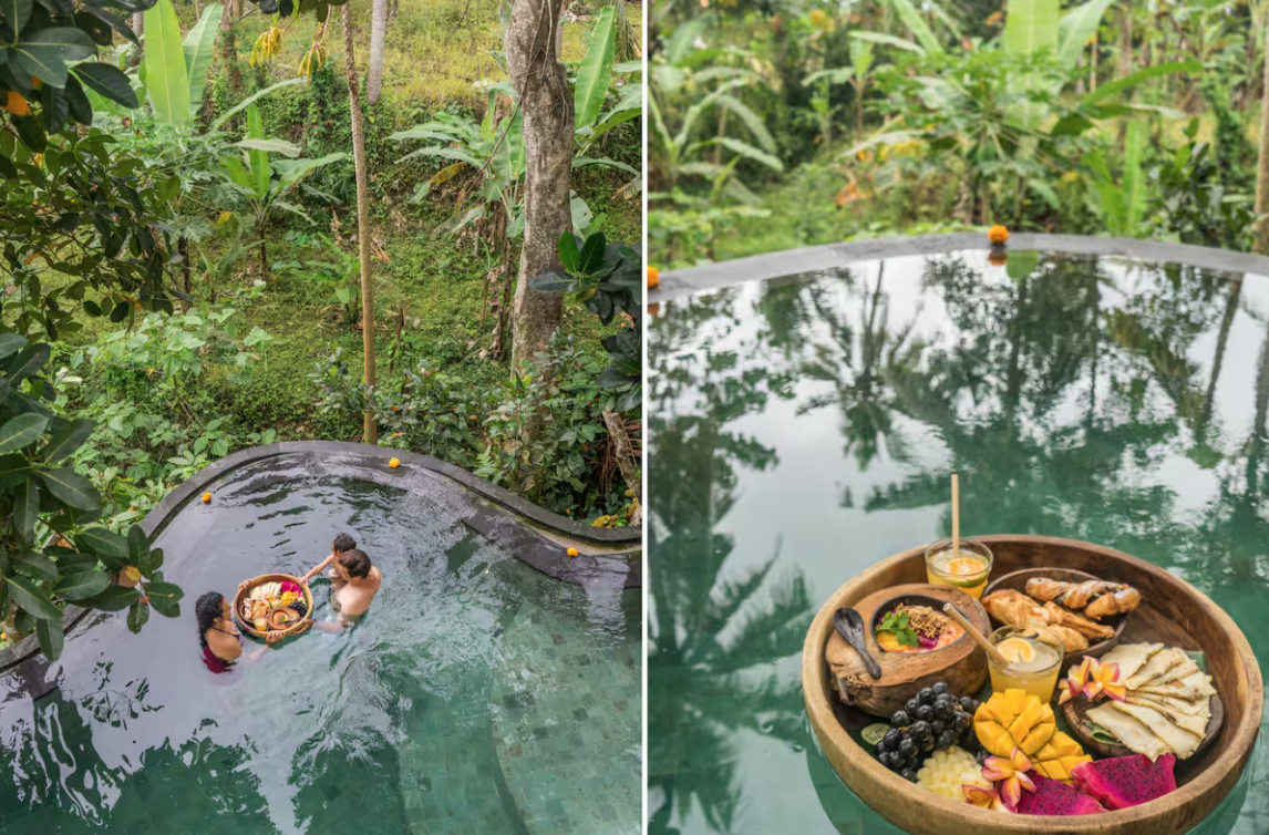 A tropical outdoor pool with three people enjoying a bath, surrounded by lush green vegetation. The other side shows a tray of breakfast foods including fruits, pastries, and drinks floating on the pool's water.