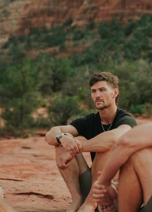 A young man sitting outdoors on a rocky surface with a desert landscape and green trees in the background, wearing a black t-shirt and gray shorts.