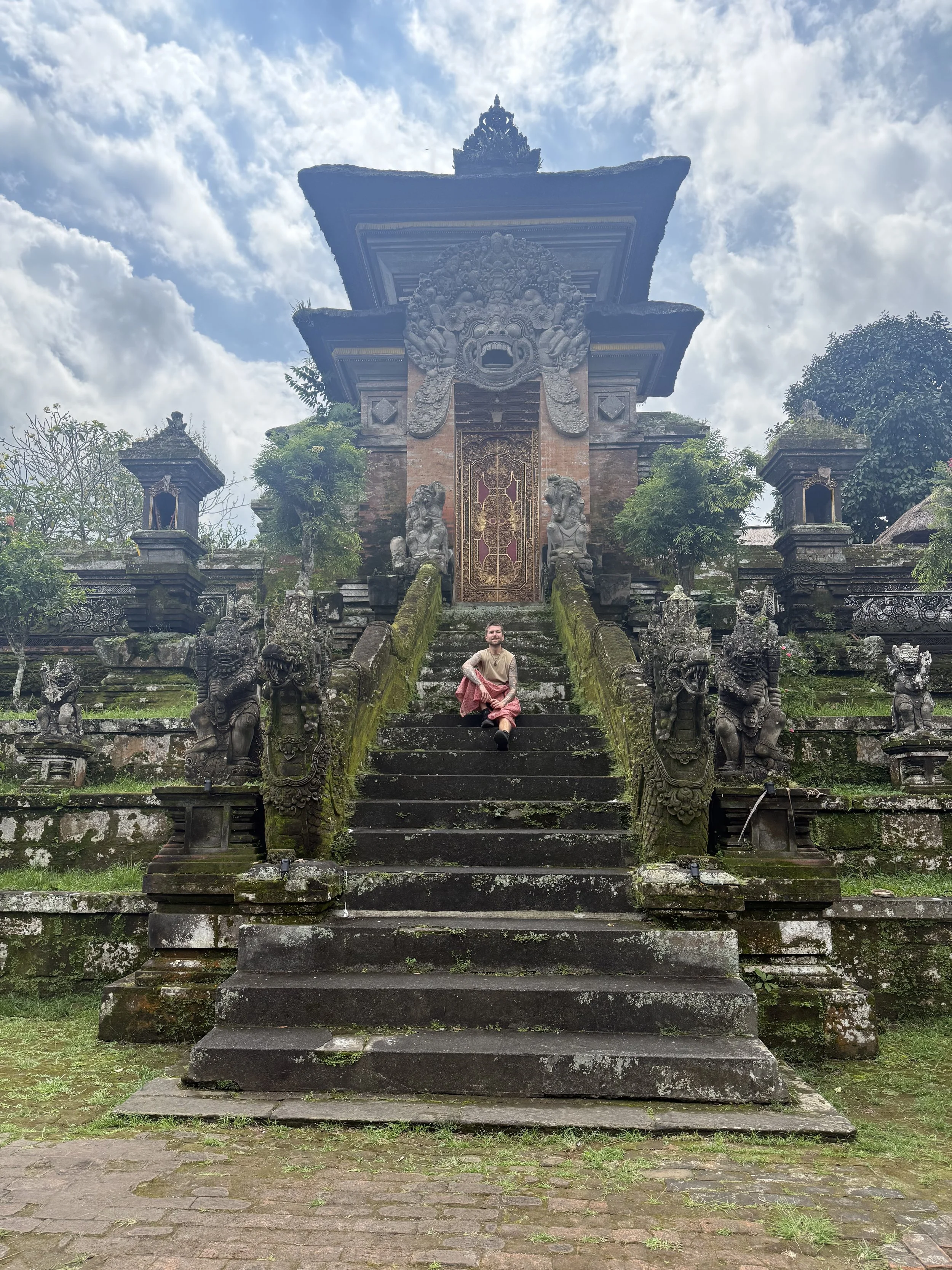 A person sitting on moss-covered stone stairs leading to an ornate traditional temple entrance with carved stone statues of lions and mythical creatures, surrounded by greenery under a cloudy sky.