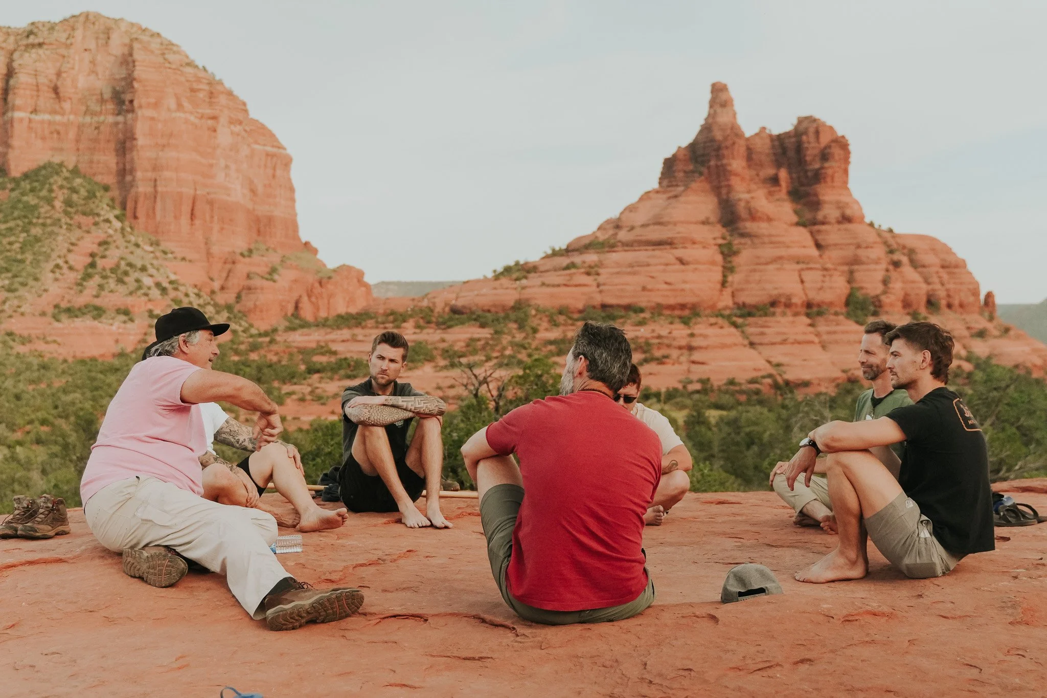 Group of people sitting cross-legged on a red rock plateau in a circle, with red sandstone mountains in the background during daytime.