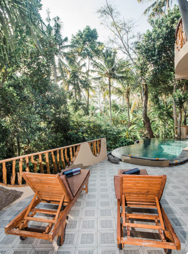 Balcony in a tropical setting with two wooden lounge chairs, a small pool, and lush green trees.