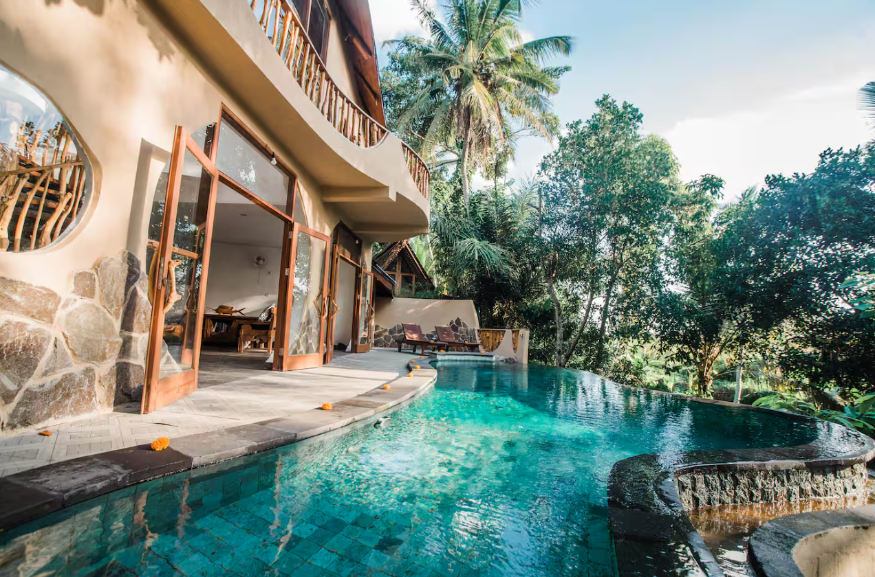 A tropical vacation home with open glass doors, overlooking a swimming pool surrounded by lush greenery and tall palm trees.