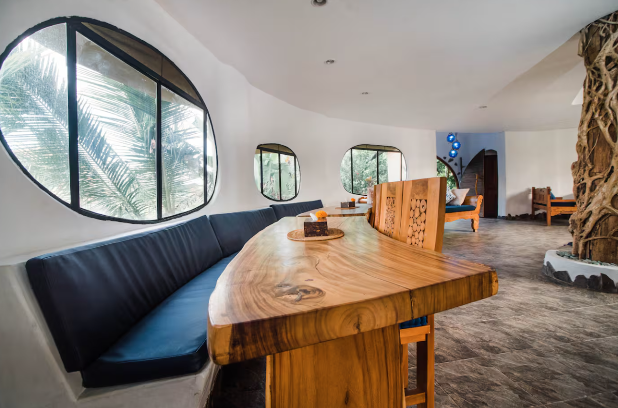 Interior of a room with curved white walls and large, uniquely shaped windows showing greenery outside. There is a long wooden table with a black cushioned bench along one side, and small decorative items on the table. In the background, additional w