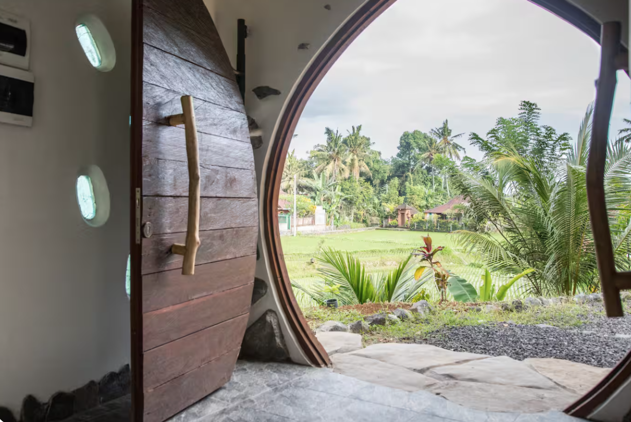 Open wooden door leading to a lush green garden with palm trees and tropical plants, visible through a round window.