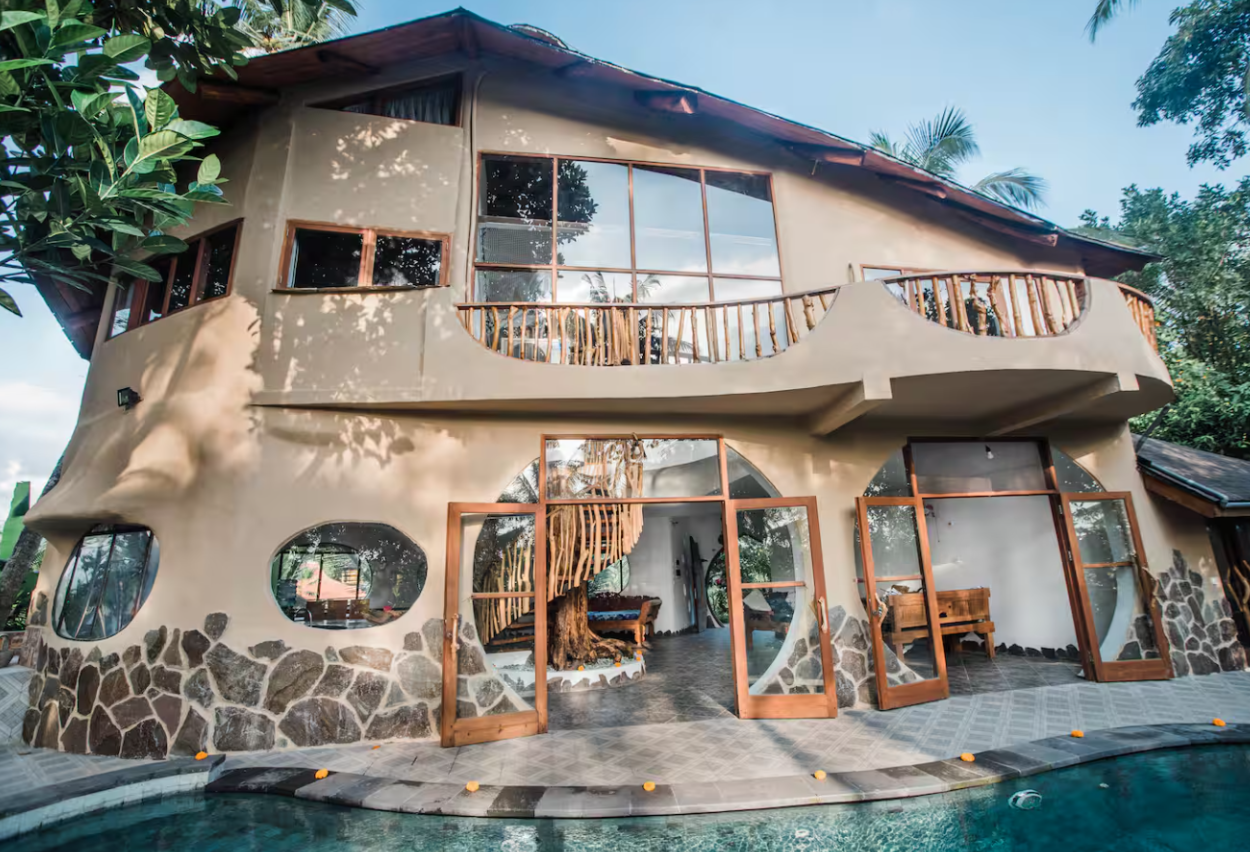 A modern, multi-story house with rounded walls and large, irregular shaped windows. The exterior features stone and stucco finishes, with a small balcony on the upper level. The house is surrounded by lush trees and greenery, and there is a swimming 