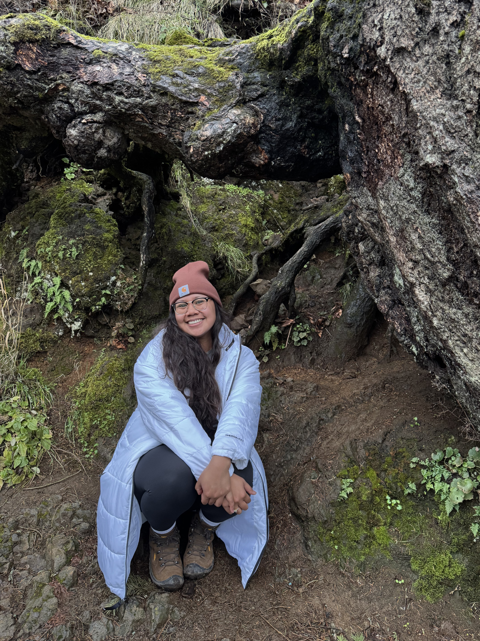 A smiling woman wearing a brown beanie, glasses, a white jacket, and hiking boots is crouching outdoors near moss-covered rocks and tree roots in a forested area.