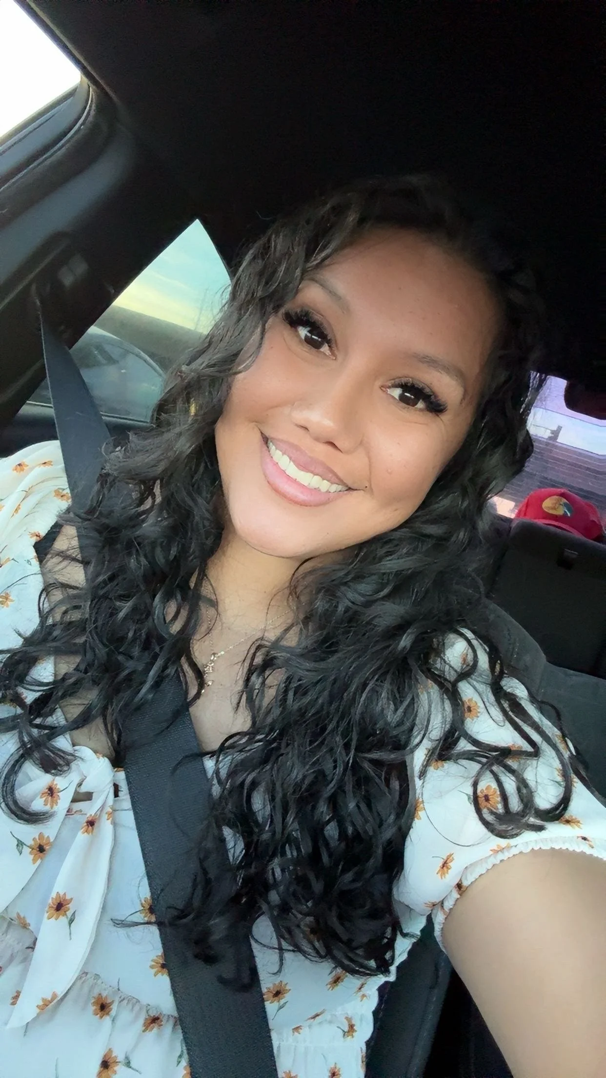 A woman with long curly black hair smiling in a car, wearing a floral top and a seatbelt.