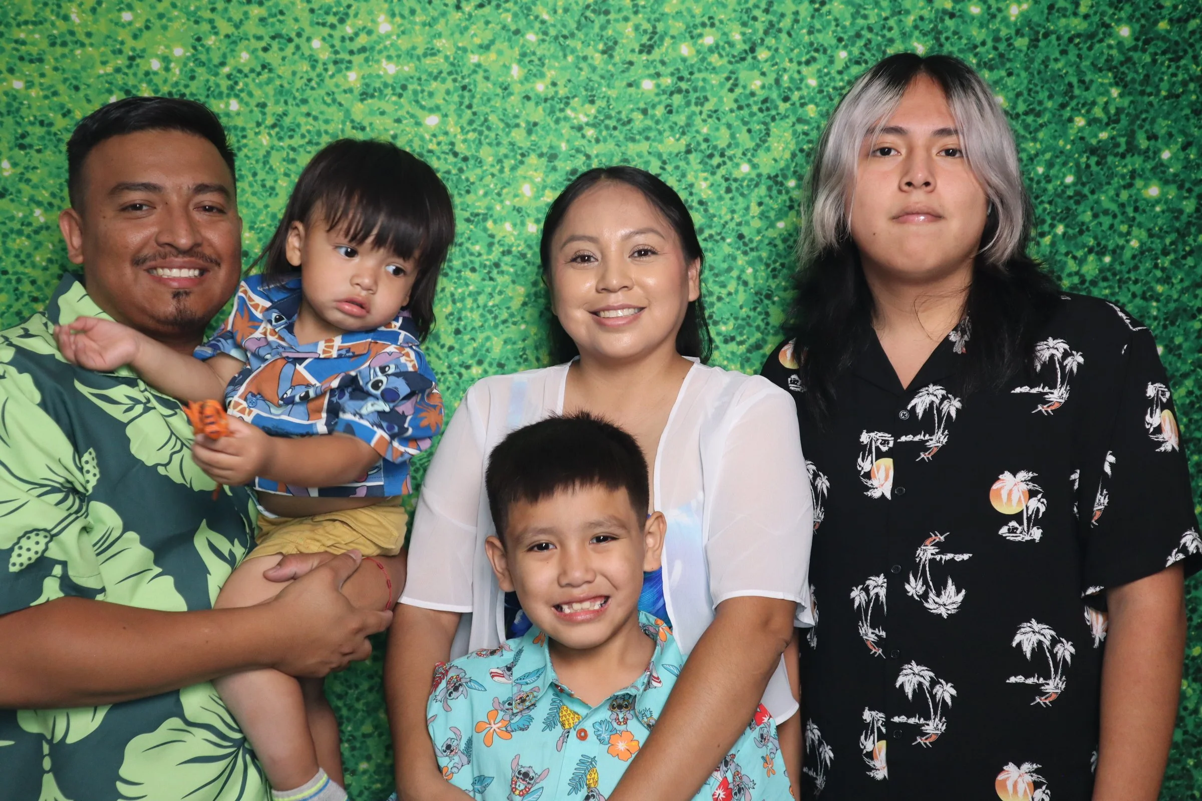 Family photo with four members, standing against a green, glittery background. From left: a man holding a young girl, a woman, and a teenage boy. All are smiling, except the girl who appears neutral.