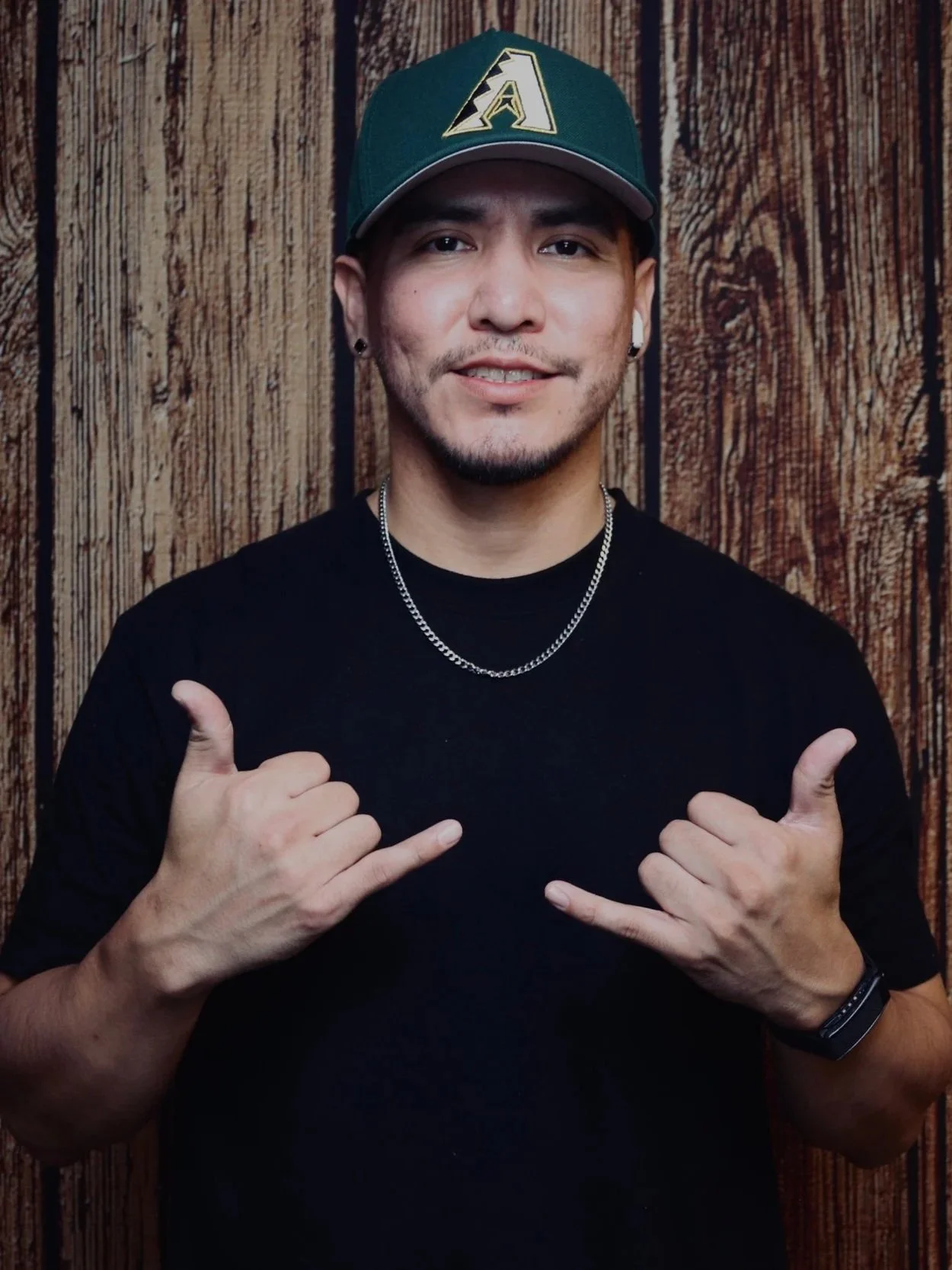 A young man with a baseball cap, earrings, and a necklace, making a shaka sign with both hands in front of a wooden background.