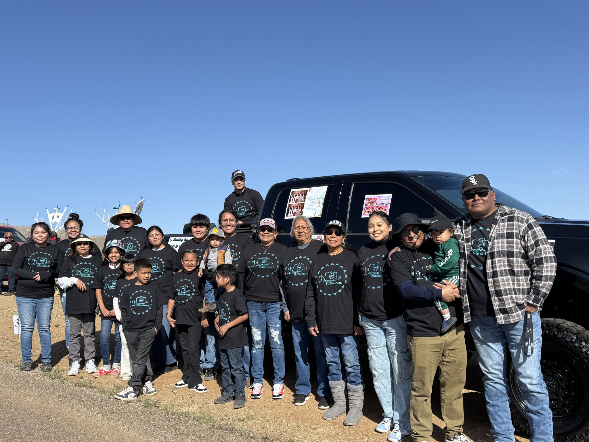 A group of people standing outdoors in front of a black vehicle, all wearing matching black shirts with a design on the front, smiling for the photo on a sunny day.
