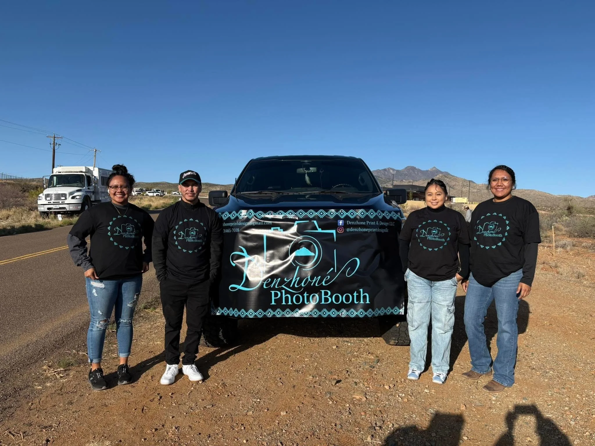 Group of four women standing next to a black vehicle with a banner reading 'Denzhône PhotoBooth' outdoors on a clear, sunny day with mountains in the background.
