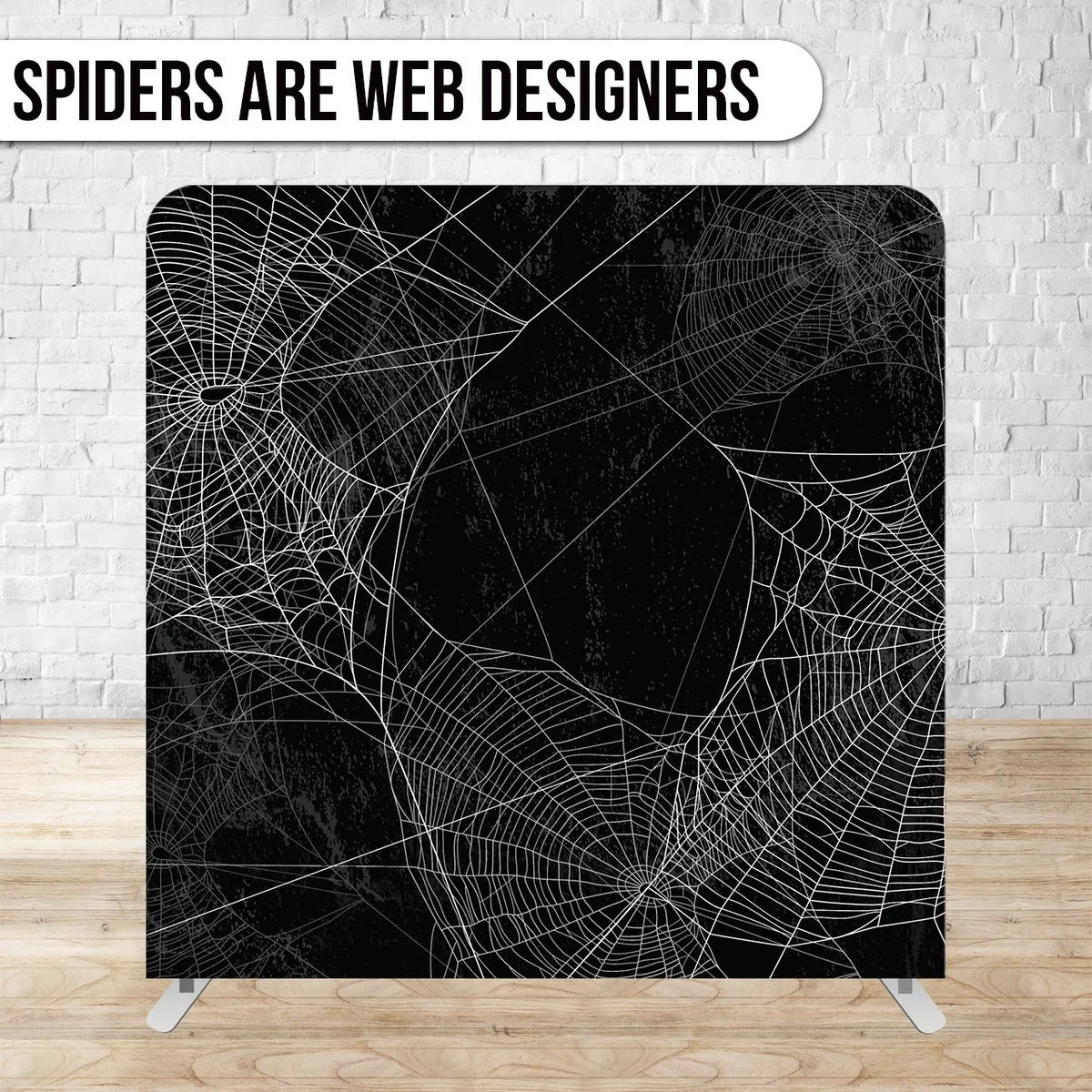 Black speaker with white spider webs design, placed on a wooden floor against a white brick wall. Text above reads "Spiders are web designers."