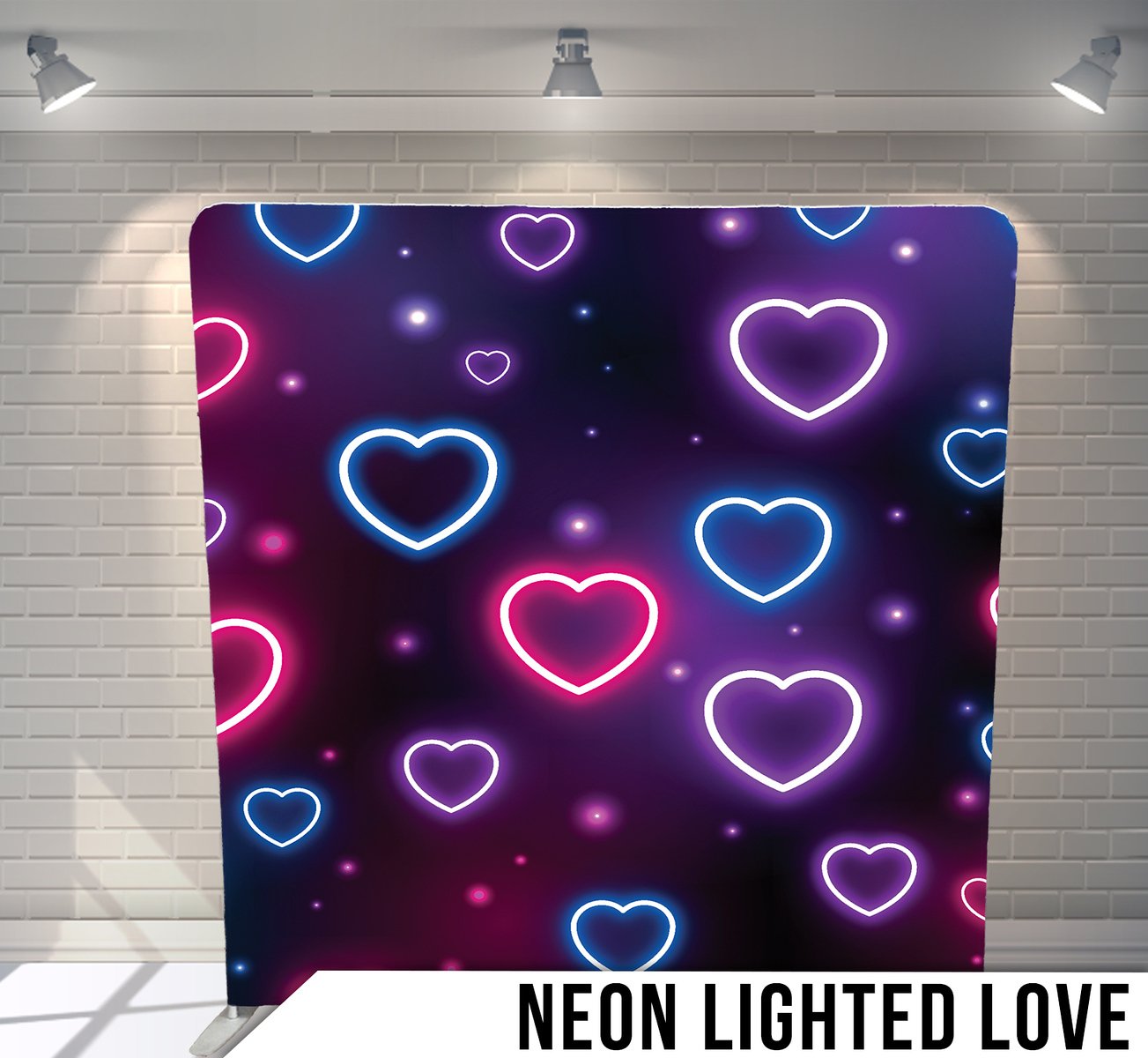 A neon-themed backdrop with glowing pink and blue heart outlines on a dark background, labeled 'Neon Lit Love' at the bottom, illuminated by overhead spotlights, set against a white brick wall.