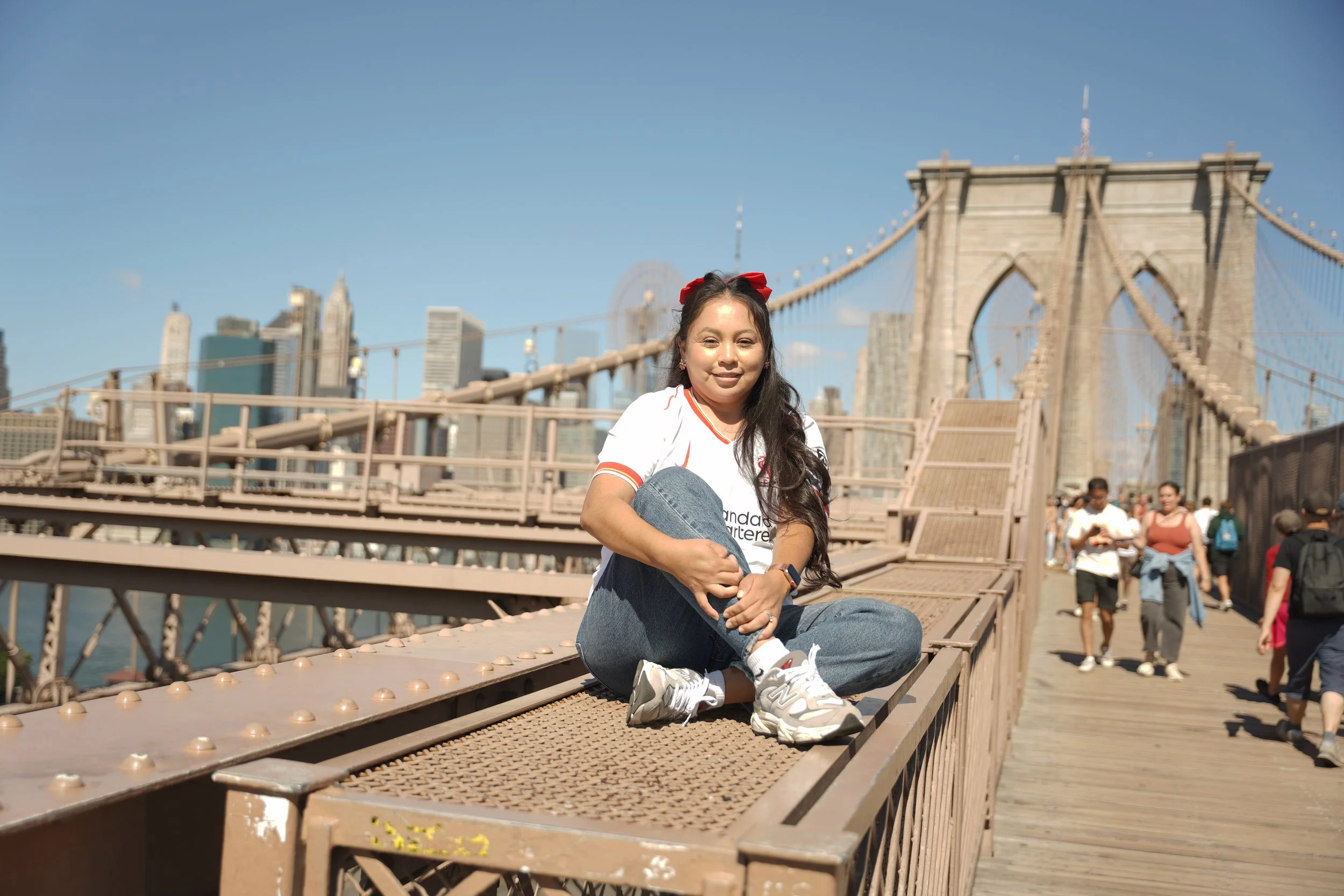 Young woman sitting cross-legged on the wooden pathway of the Brooklyn Bridge in New York City, with the Manhattan skyline in the background during daytime.
