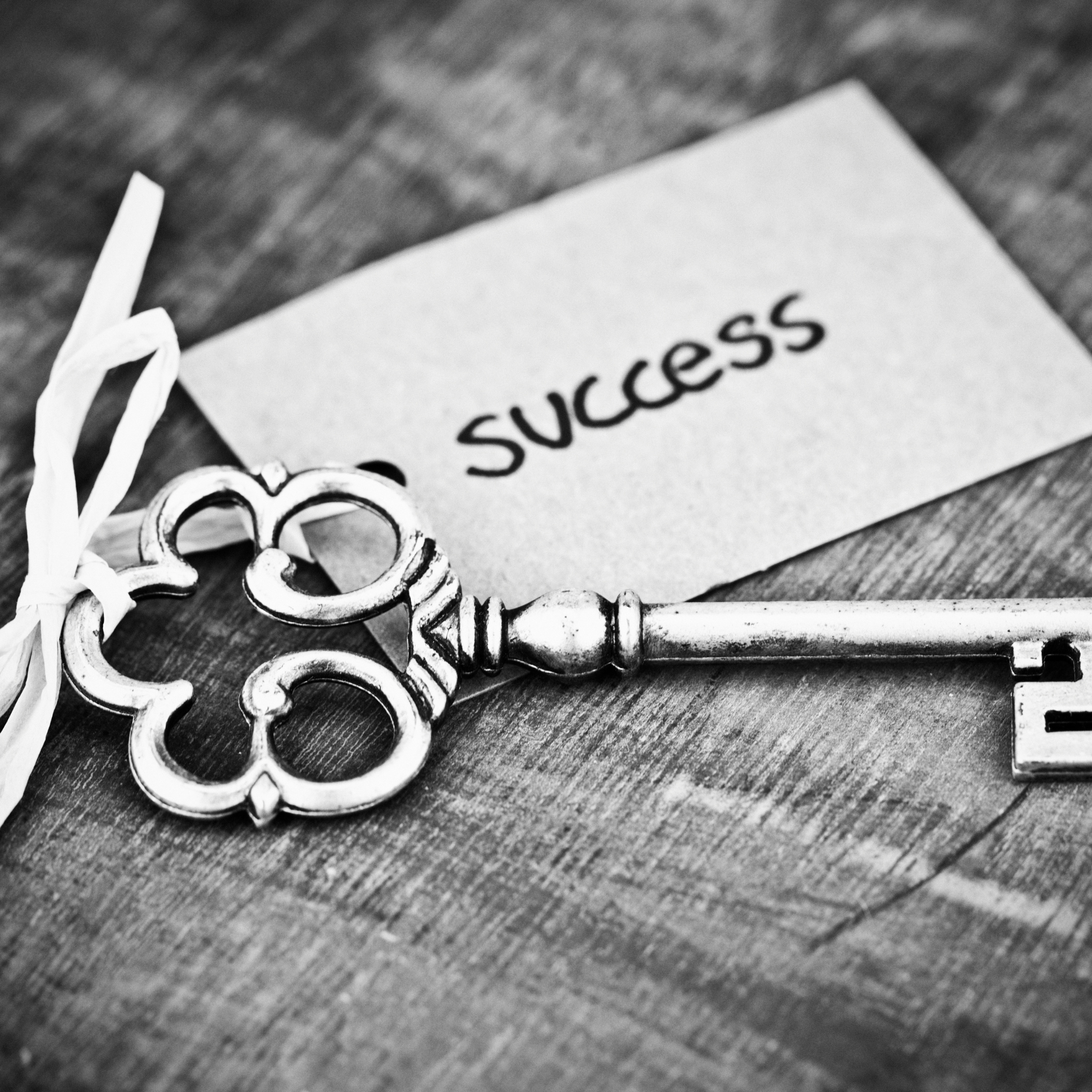 A close-up of a decorative metal key resting on a wooden surface next to a paper with the word 'success' written on it. The photo is in black and white.