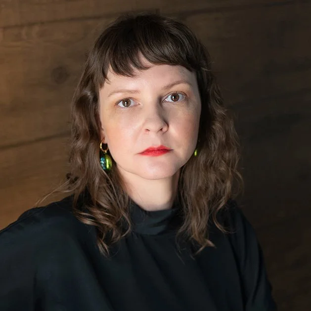 A woman with light skin, shoulder-length wavy brown hair, and red lipstick, wearing green earrings and a black top, standing against a wooden background.