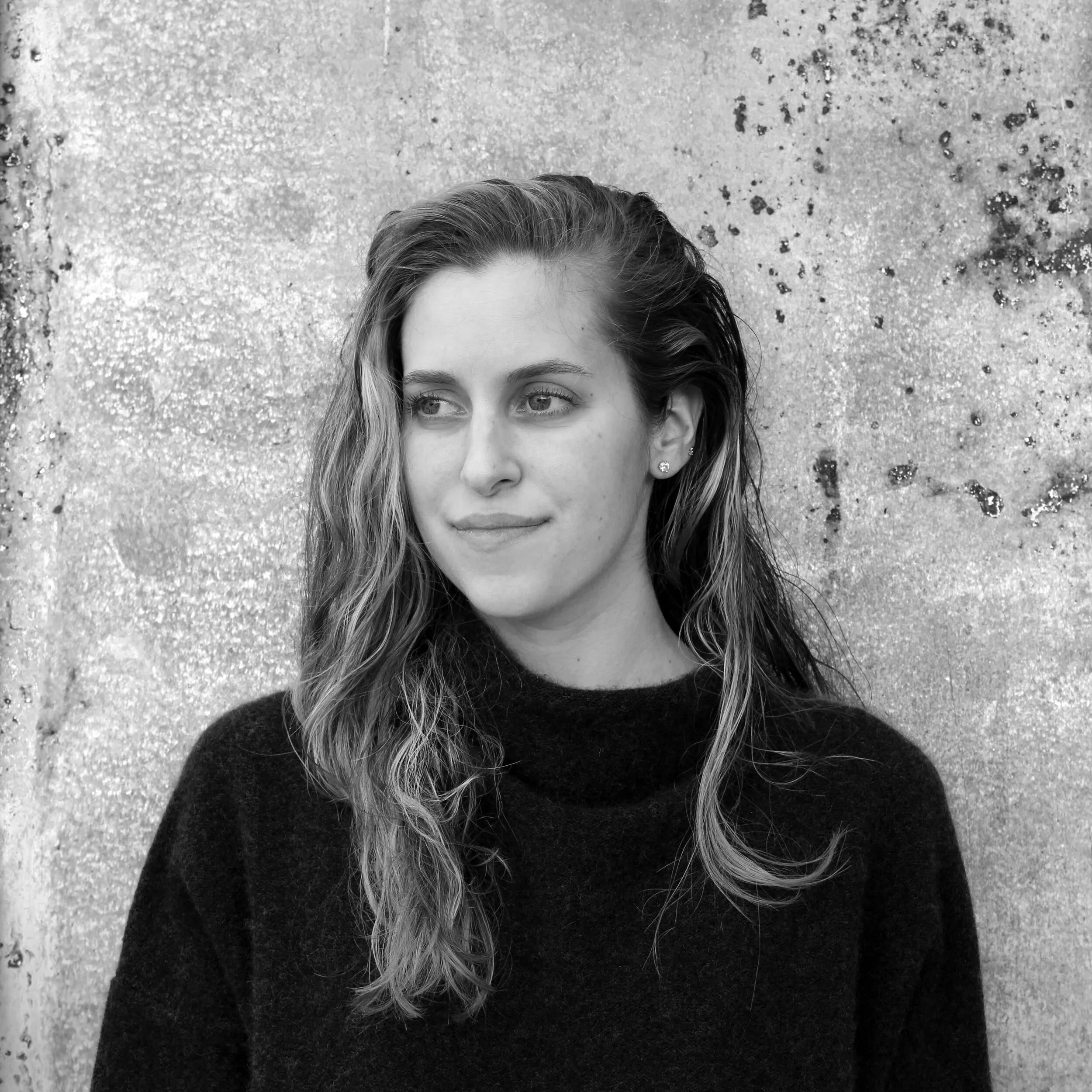 A young woman with wavy hair and stud earrings looking to her left in front of a textured wall.