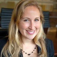 A young woman with blonde hair smiling, wearing a black blazer and a beaded necklace.
