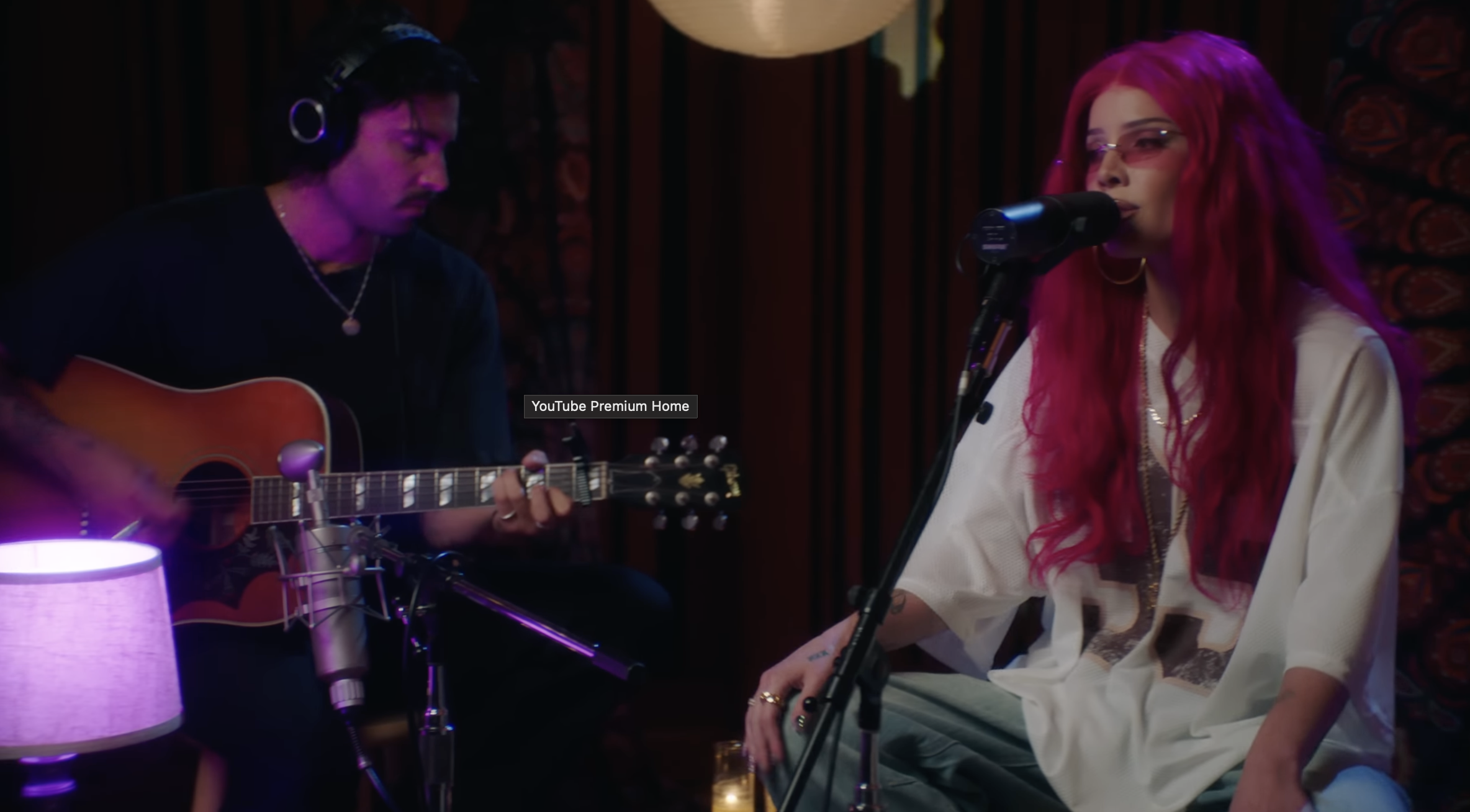 Halsey singing with long pink hair singing into a microphone, and a man with dark hair playing an acoustic guitar in a dimly lit room.