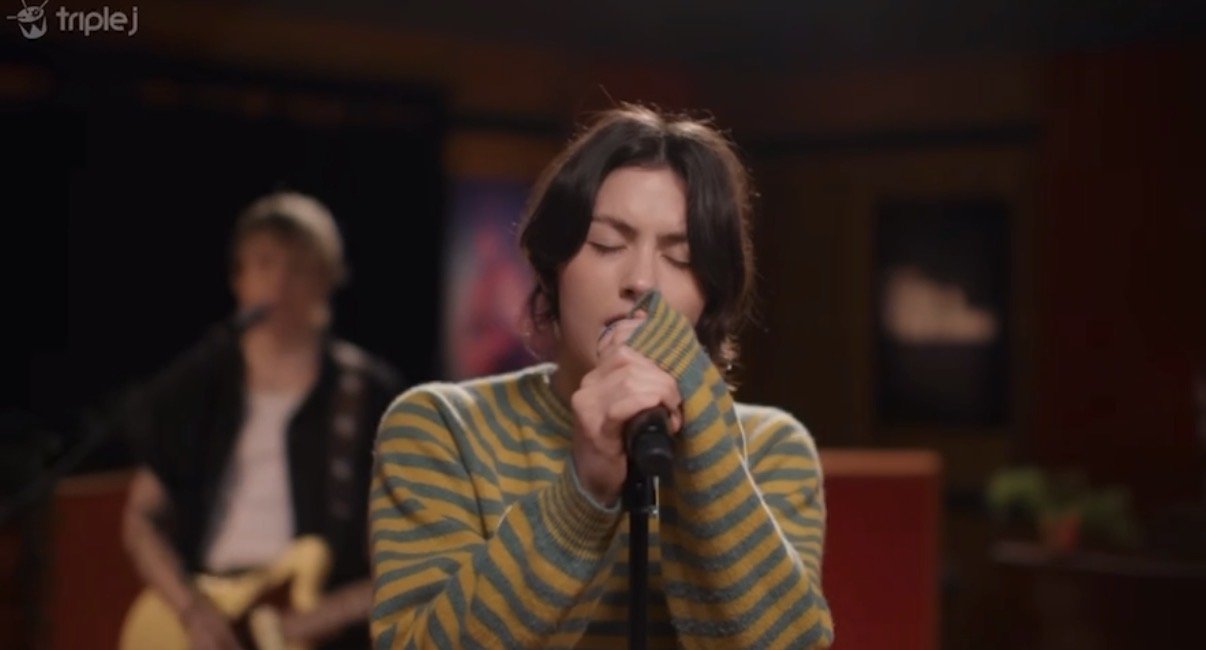 Gracie Abrams in a striped sweater sings into a microphone with eyes closed. In the background, a man plays a guitar.