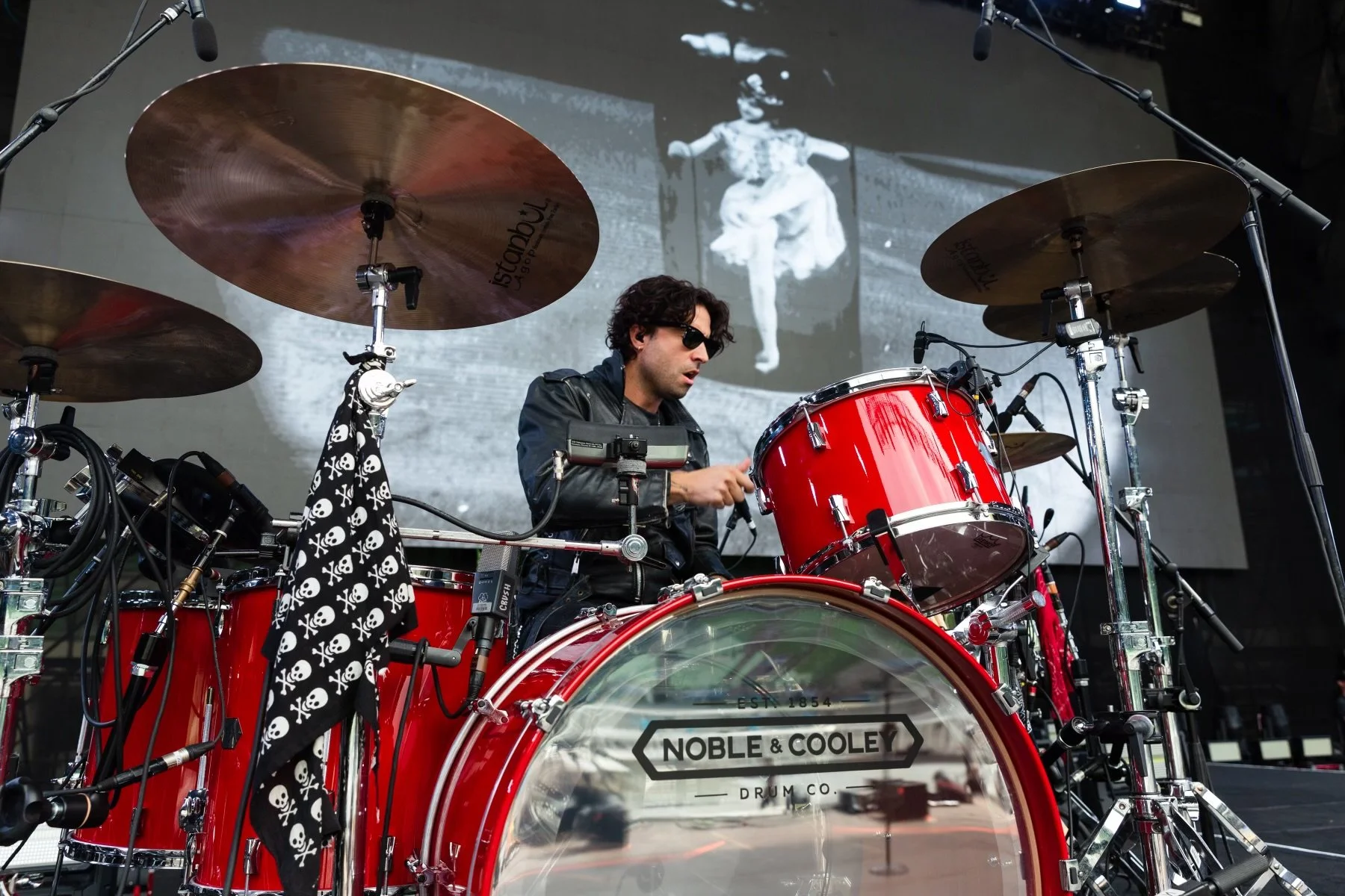 A drummer wearing sunglasses and a leather jacket playing a red drum set on stage, with a black and white skull-patterned cloth hanging from the drums and a black-and-white background image projected on a screen behind him.