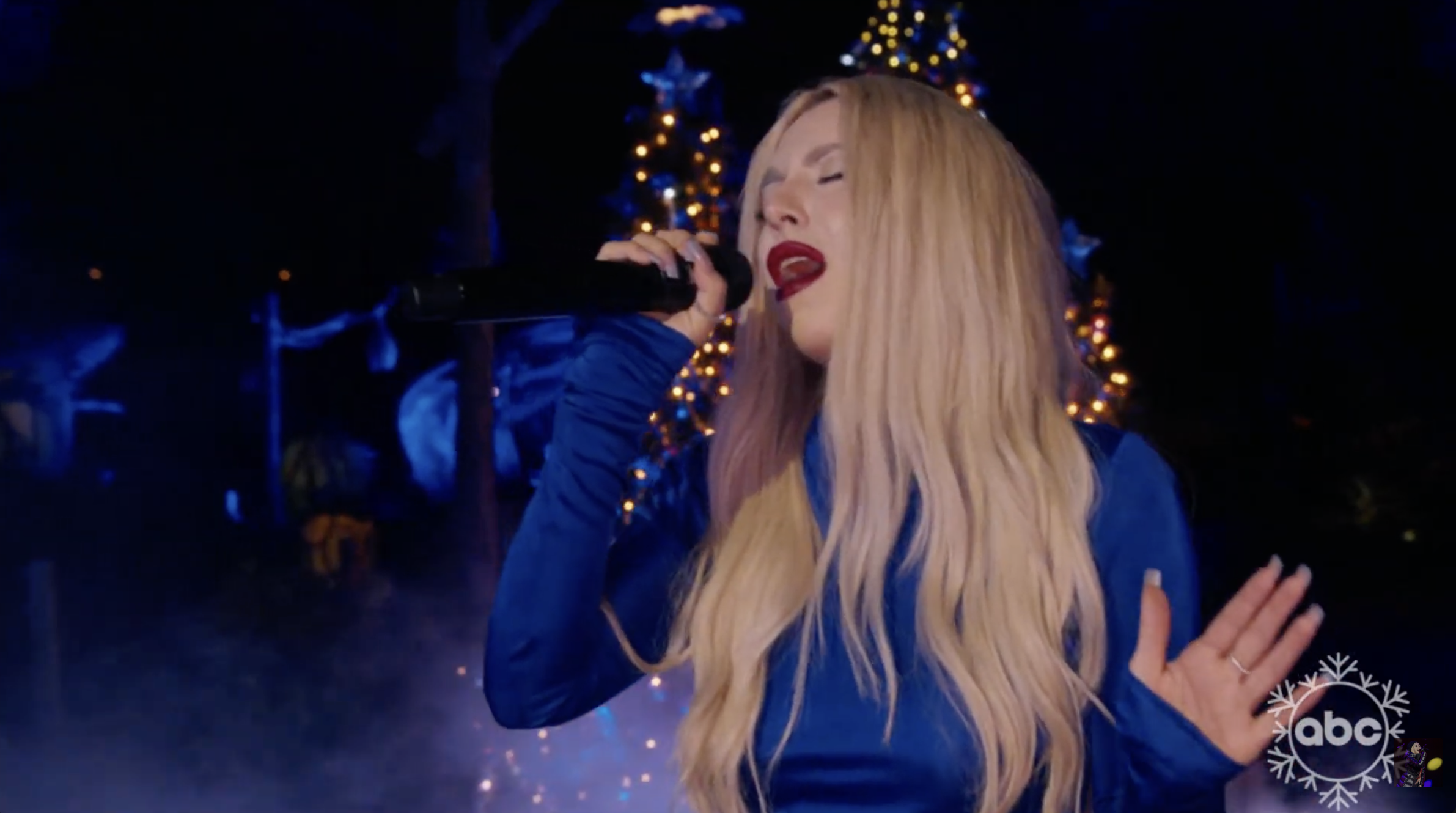 Ava Max with long blonde hair singing into a microphone on stage, with Christmas trees and festive lights in the background.