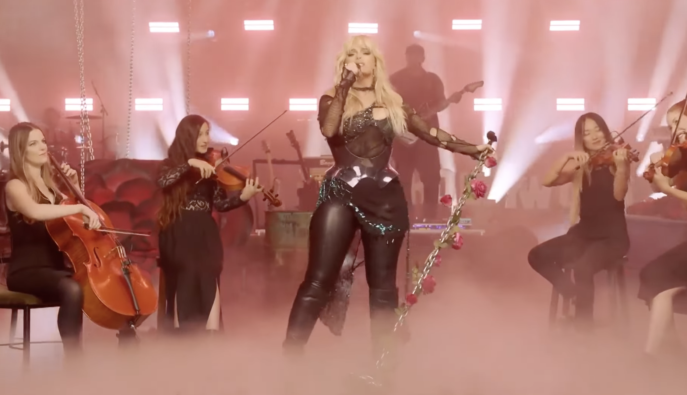Bebe Rexha in black leather pants and a sheer black top singing into a microphone surrounded by a band playing violins, guitar, and keyboard, with pink lighting and fog.