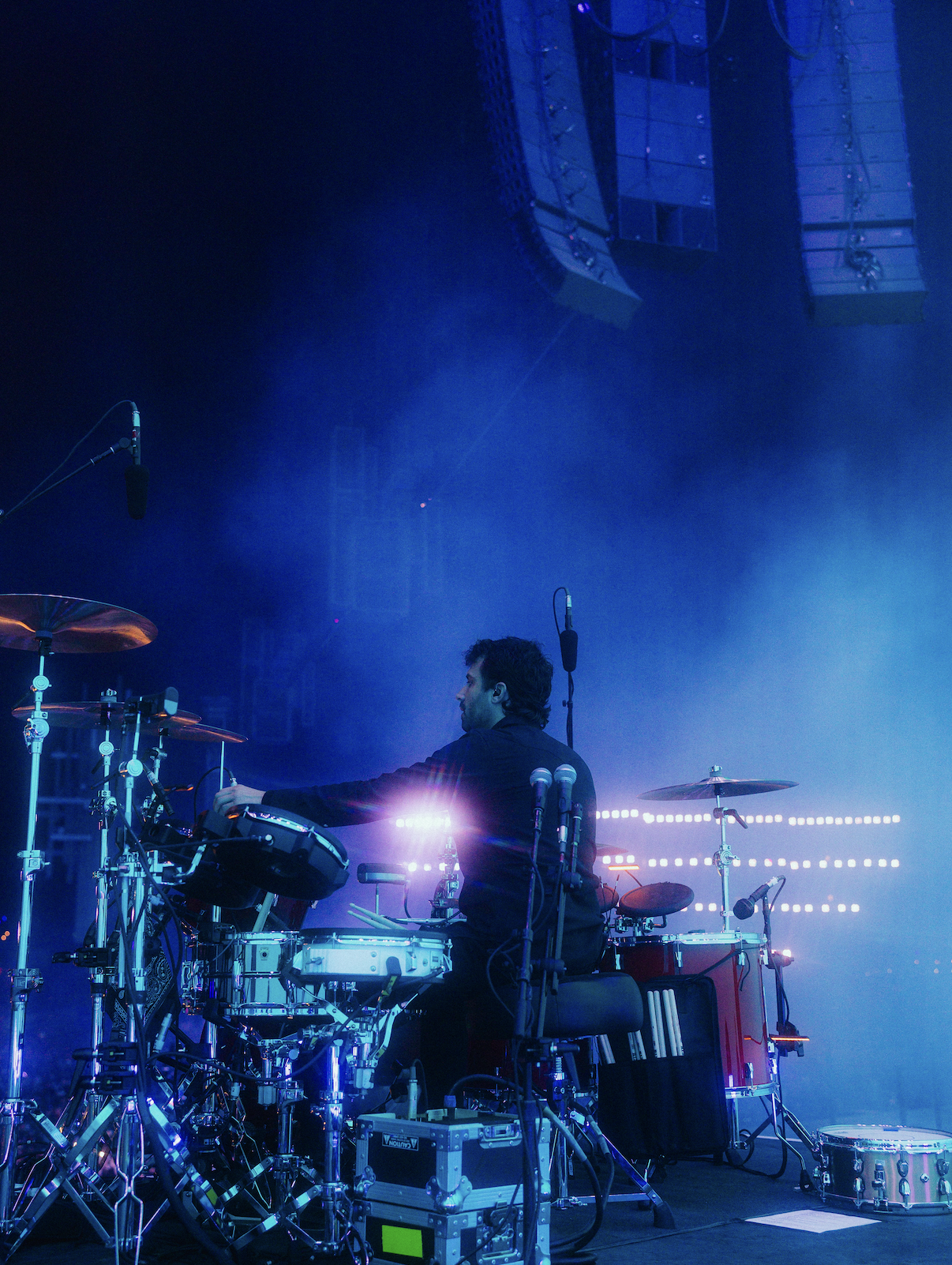 Musician performing on stage with electronic drums and equipment, surrounded by blue and purple lighting, with large speakers overhead.