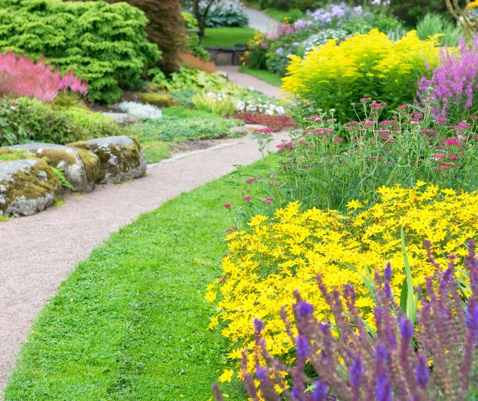 Colorful garden with a curved gravel pathway, lush green grass, and a variety of vibrant flowers including yellow, purple, and pink blooms, surrounded by trees and shrubs.